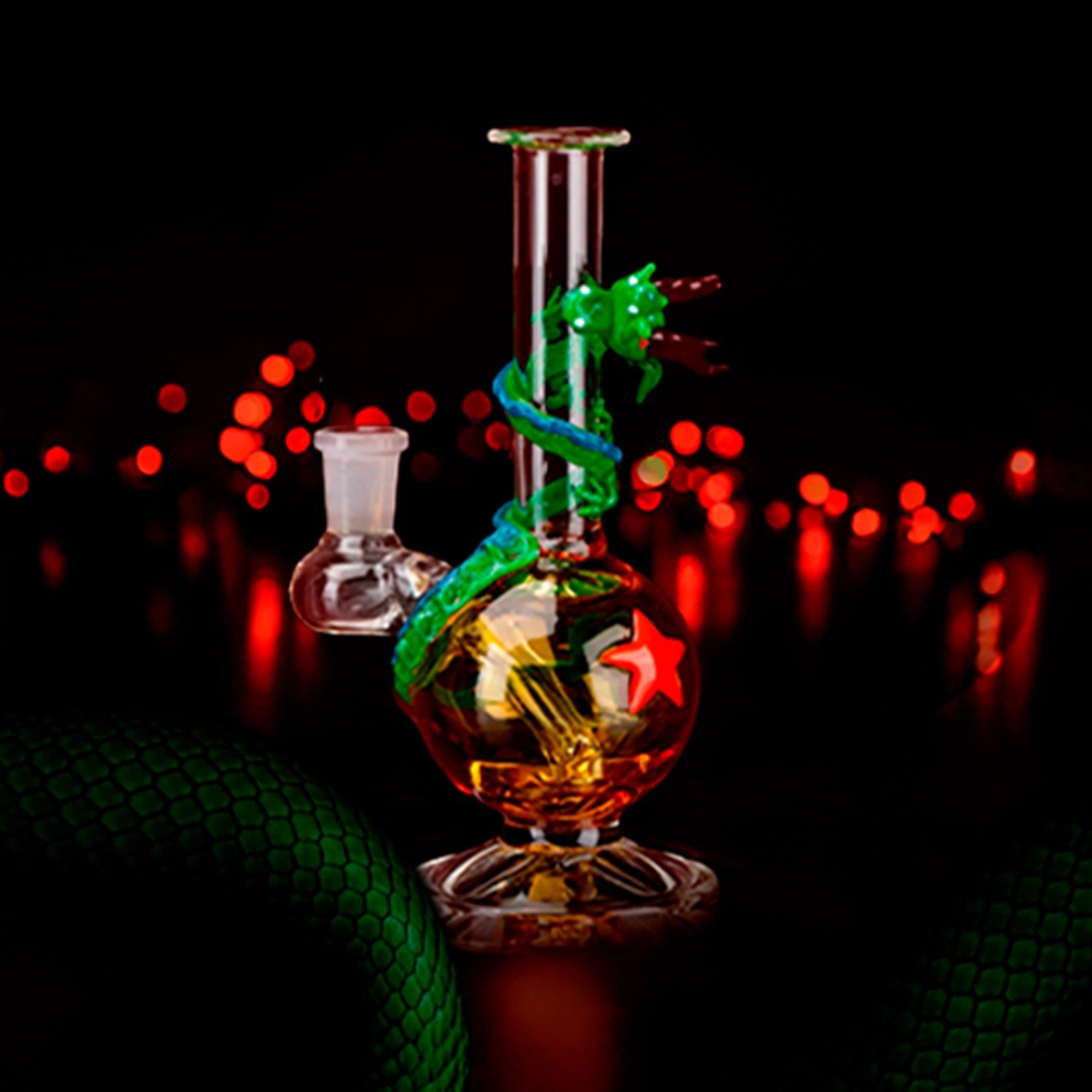 Center-front view of the Empire Glassworks Dragon Sphere Mini-Tube with a green glass dragon spiraling around a glowing amber globe against red bokeh lights