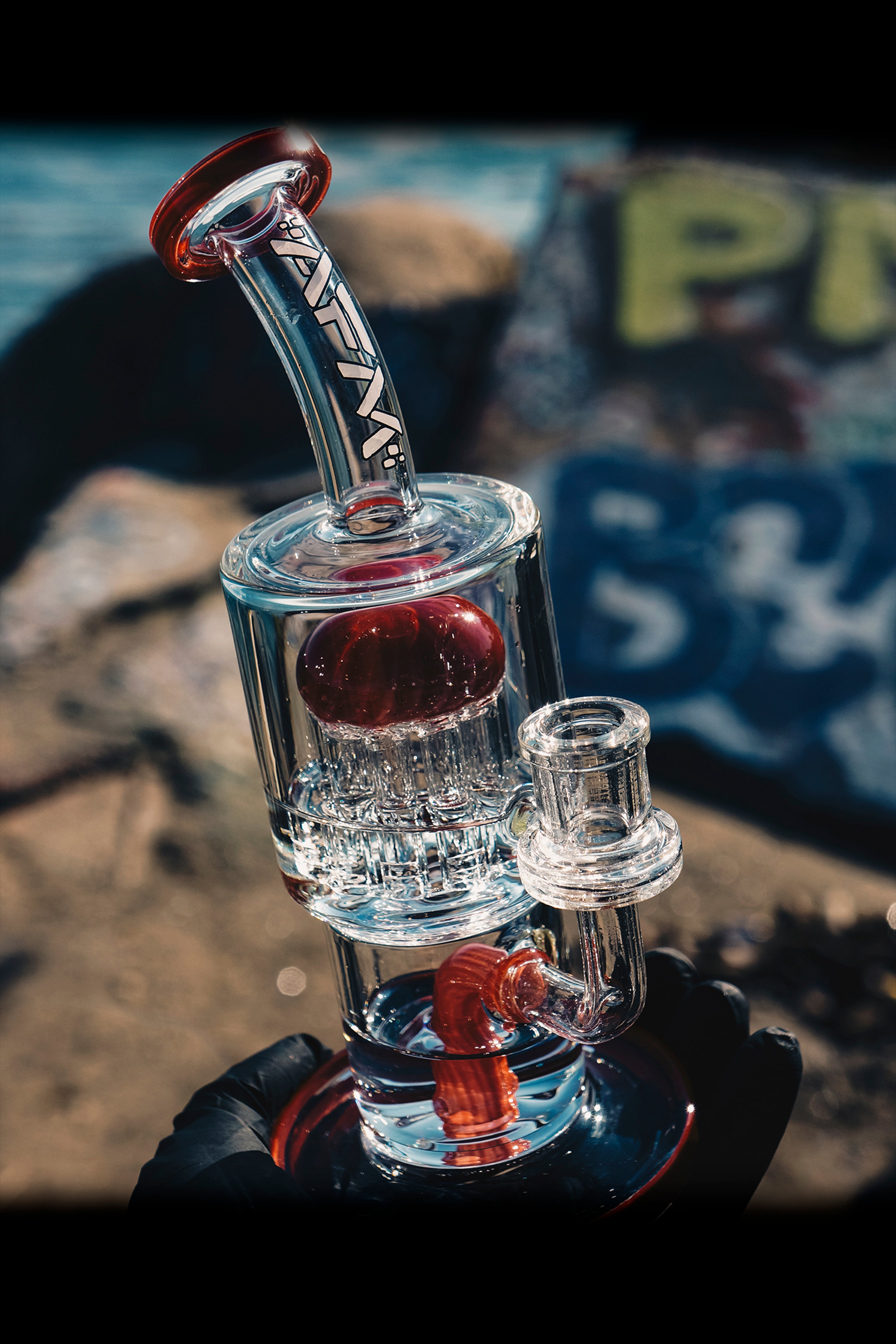 A clear glass water pipe (rig) with red accents, showcasing a complex bubbler design, sits on a black surface with a blurred background.