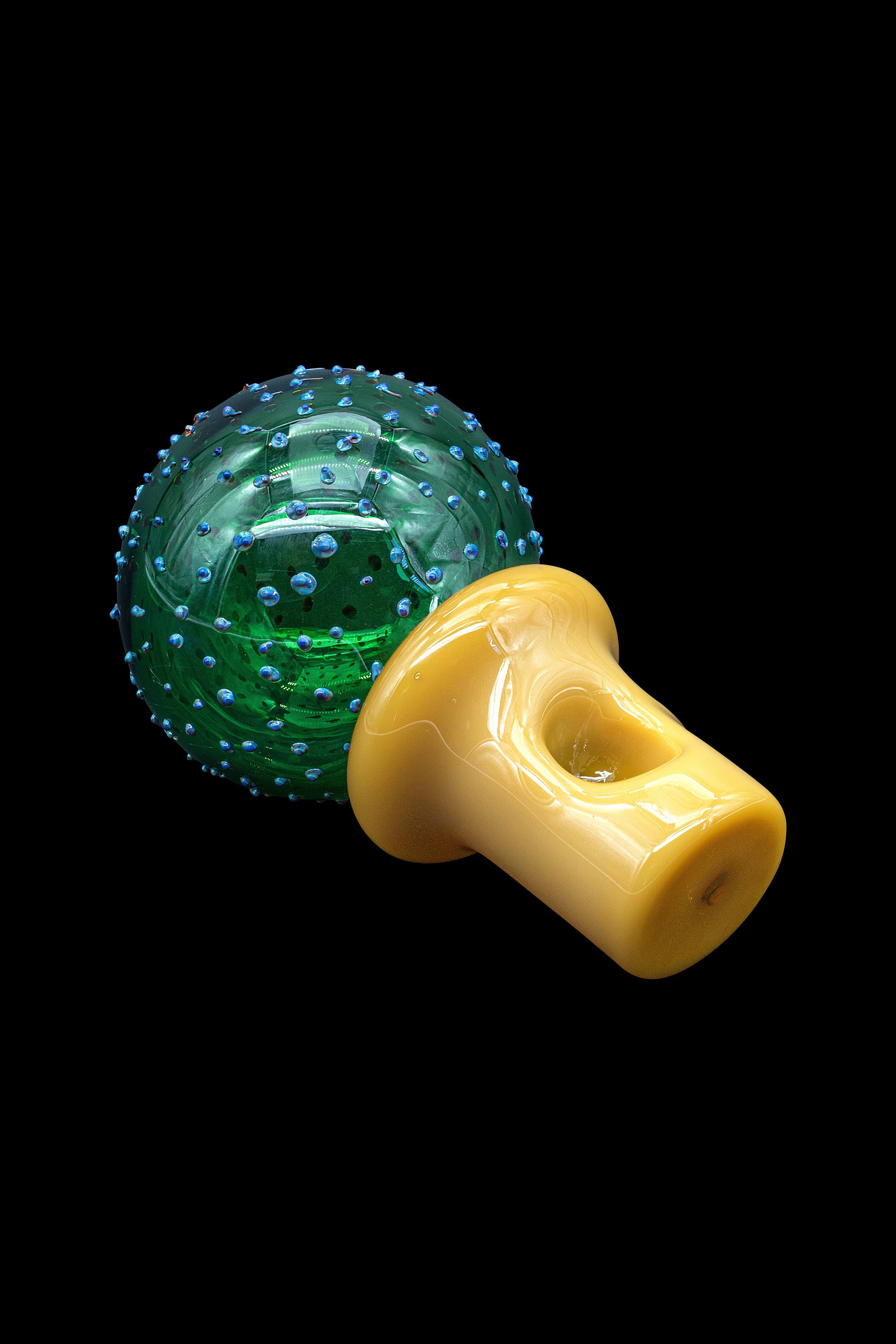 Green textured glass sphere with a yellow mushroom-shaped glass pipe resting beside it on a black background.