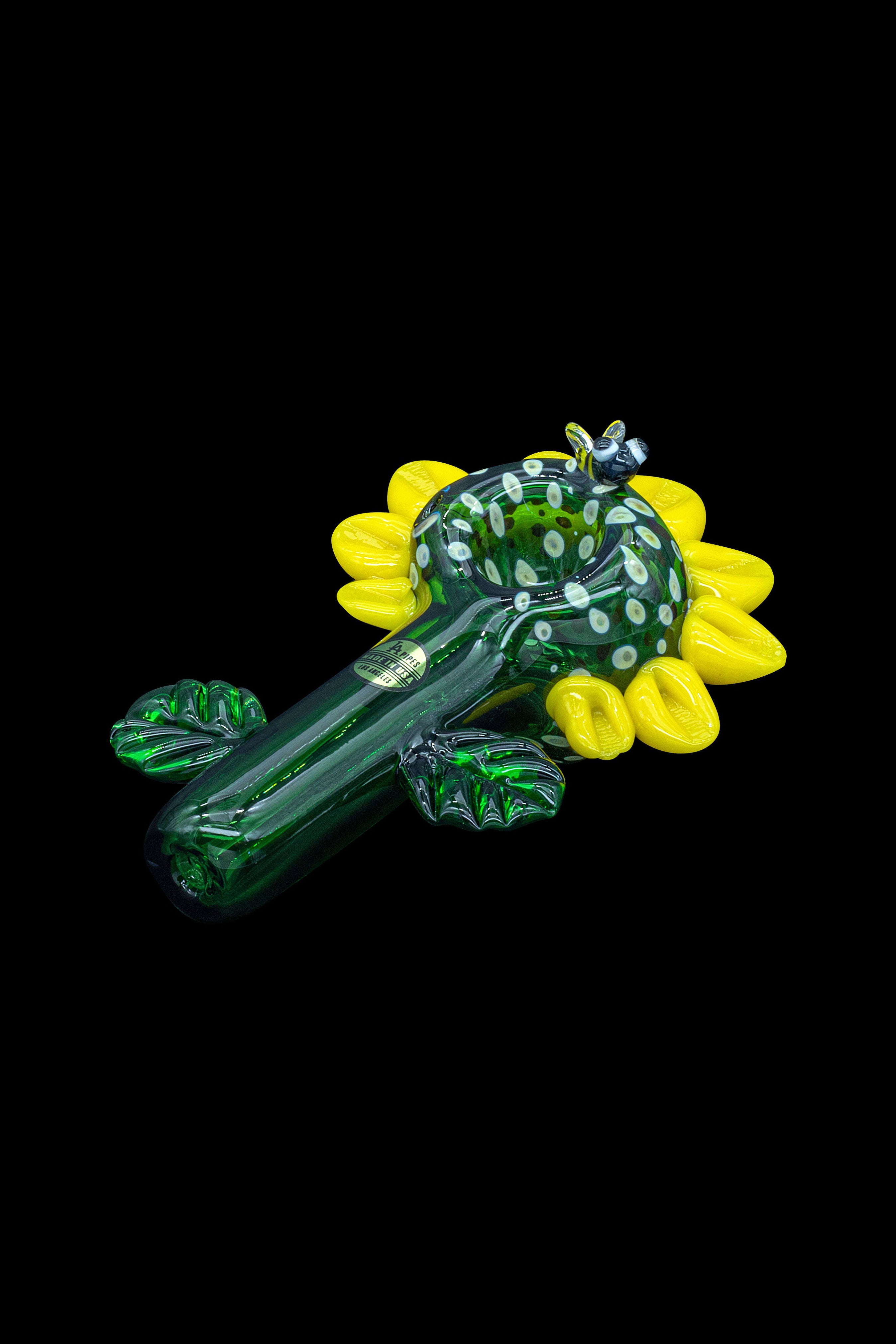 Hand-blown glass pipe shaped like a sunflower, with green and yellow hues, against a black background.