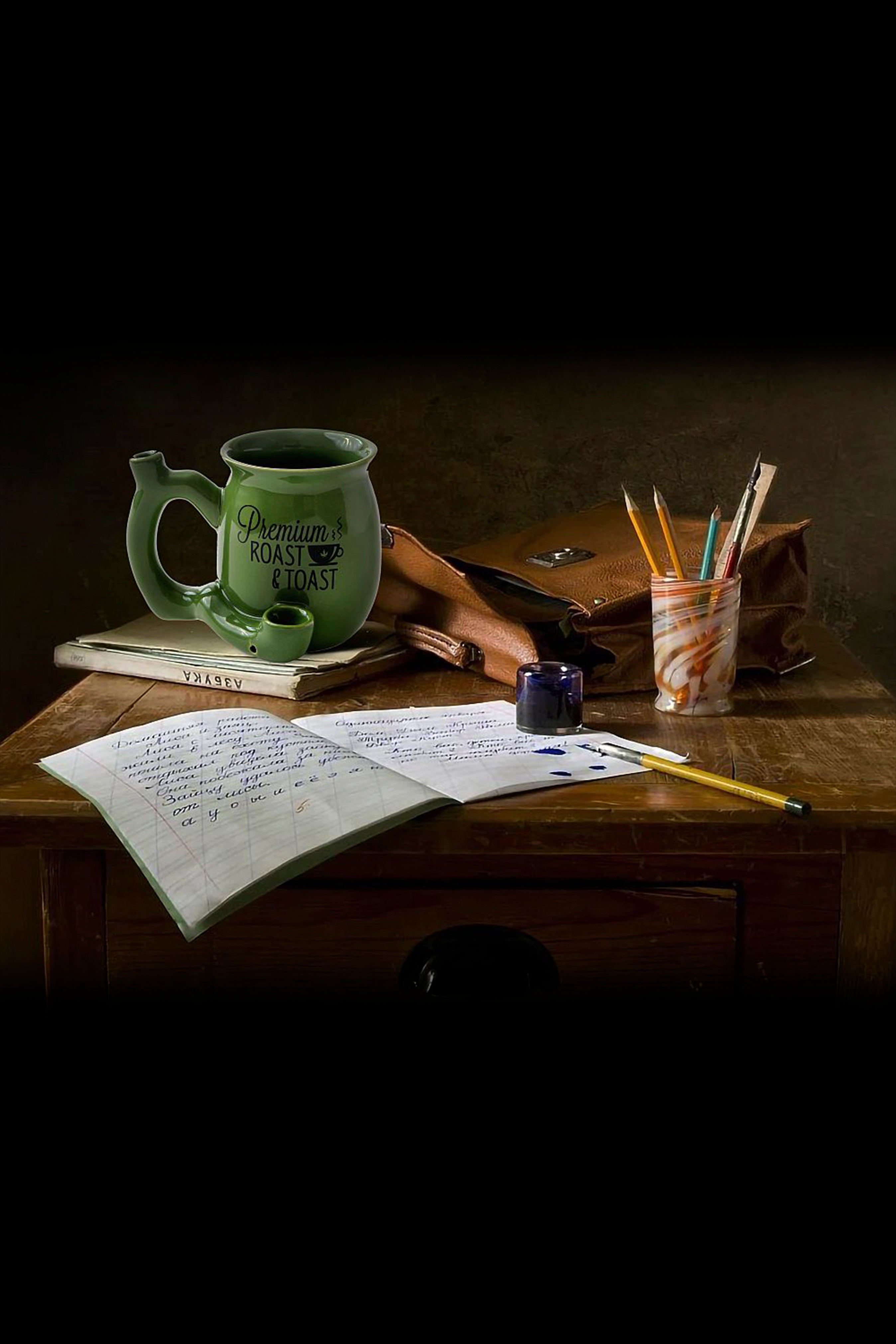 Vintage-style still life featuring a green cannabis-themed mug, open journal, writing tools, and rolling papers on a wooden desk, illuminated by warm, moody lighting.