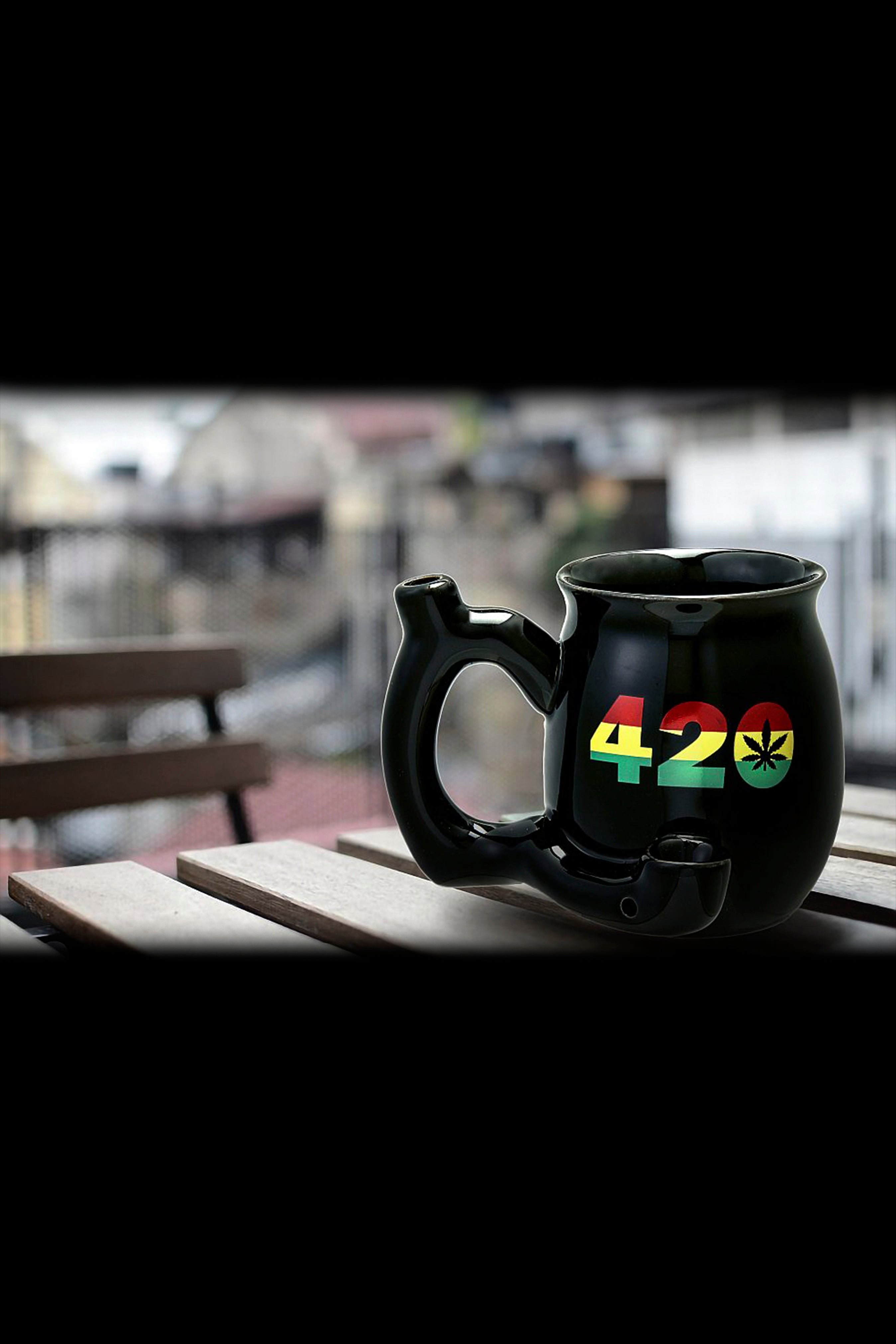 Black ceramic mug with “420” and a marijuana leaf design, sitting on a wooden table with a blurred outdoor background.