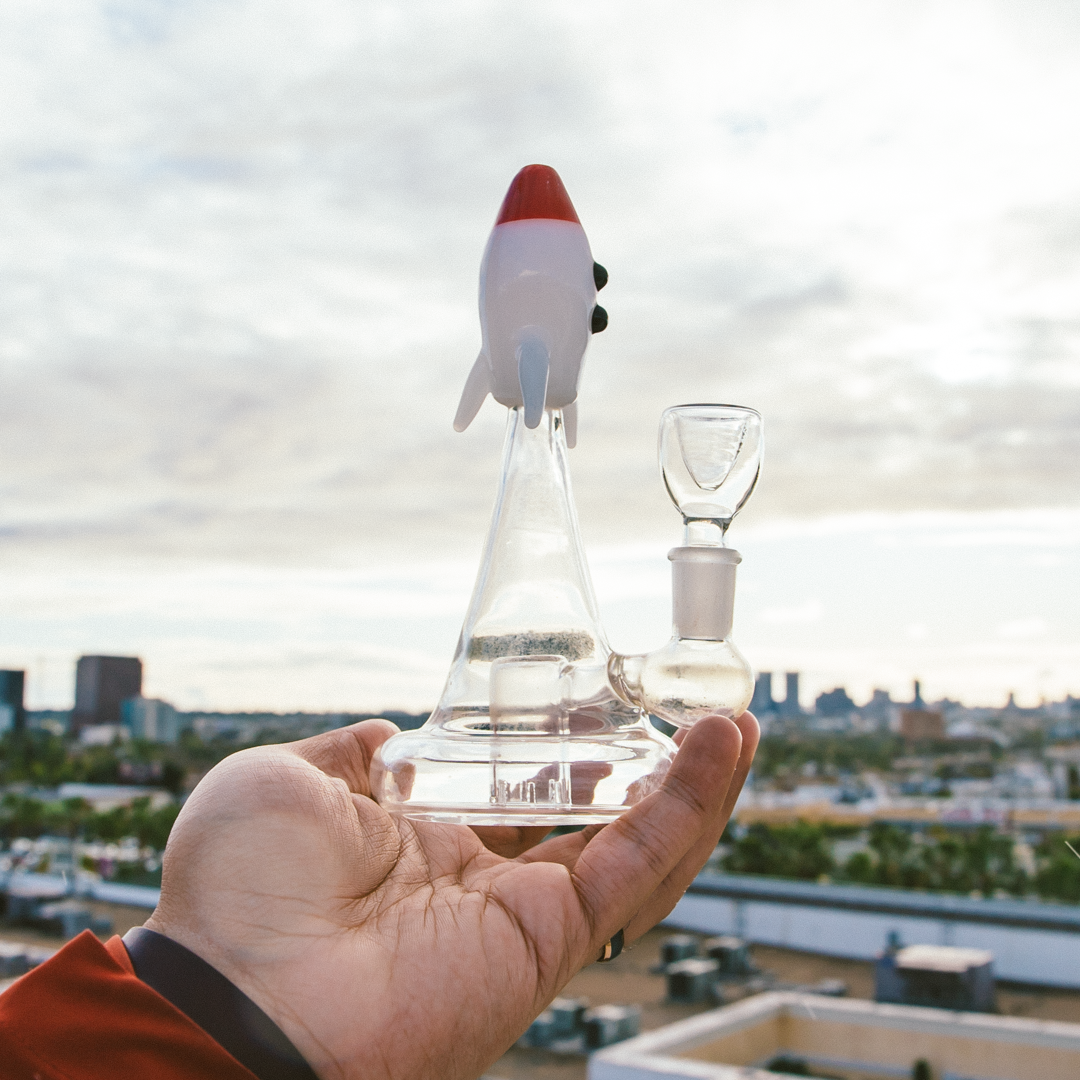 A hand holds a glass water pipe shaped like a rocket ship against a city skyline.