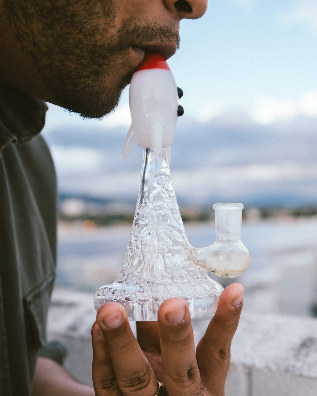 A person holds and inhales from a rocket-shaped glass water pipe, with a blurred landscape background.
