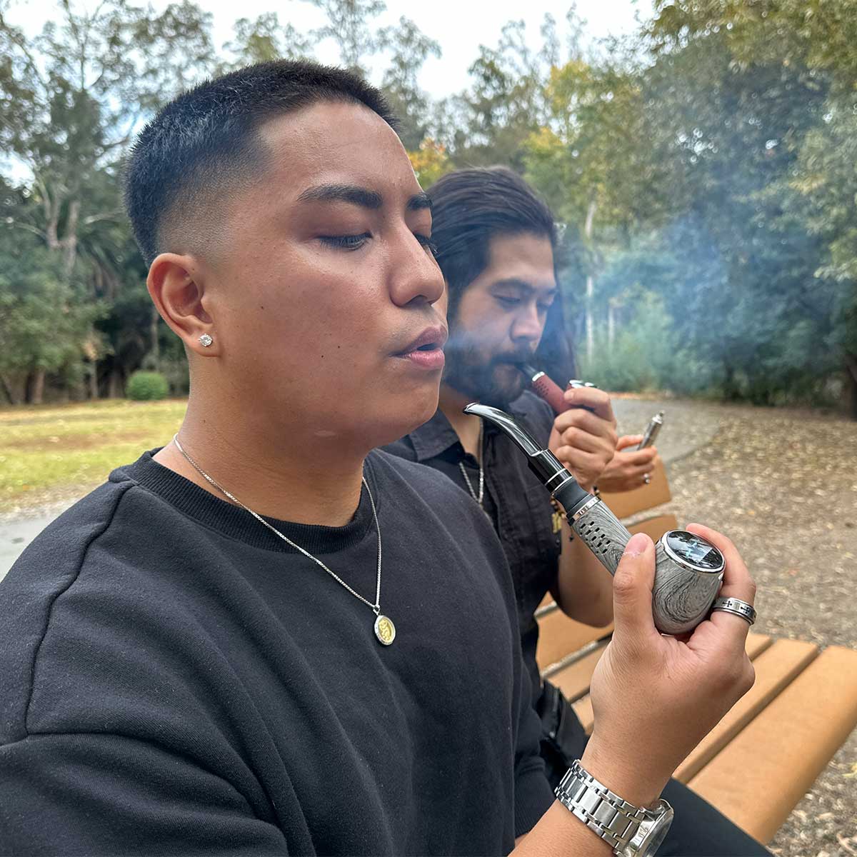 Close-up side view of a person holding and smoking the Cipher Nautilus V3 Churchwarden Stem Kit pipe outdoors on a park bench with trees and another person smoking in the background.