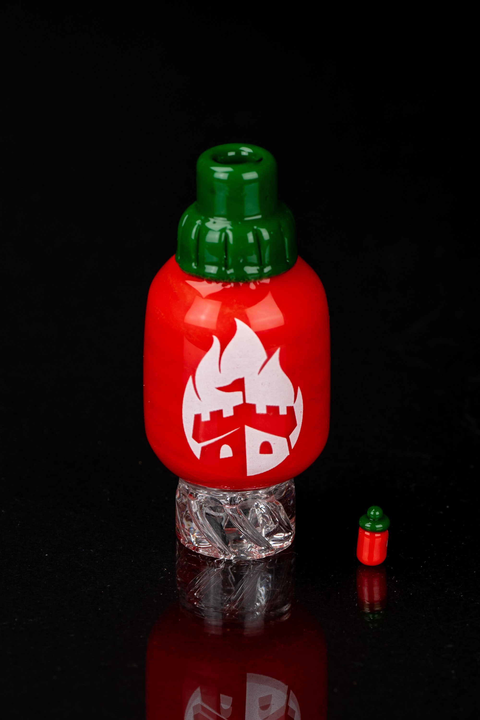 Red glass dab rig shaped like a chili pepper with a green cap, reflecting on a textured surface.