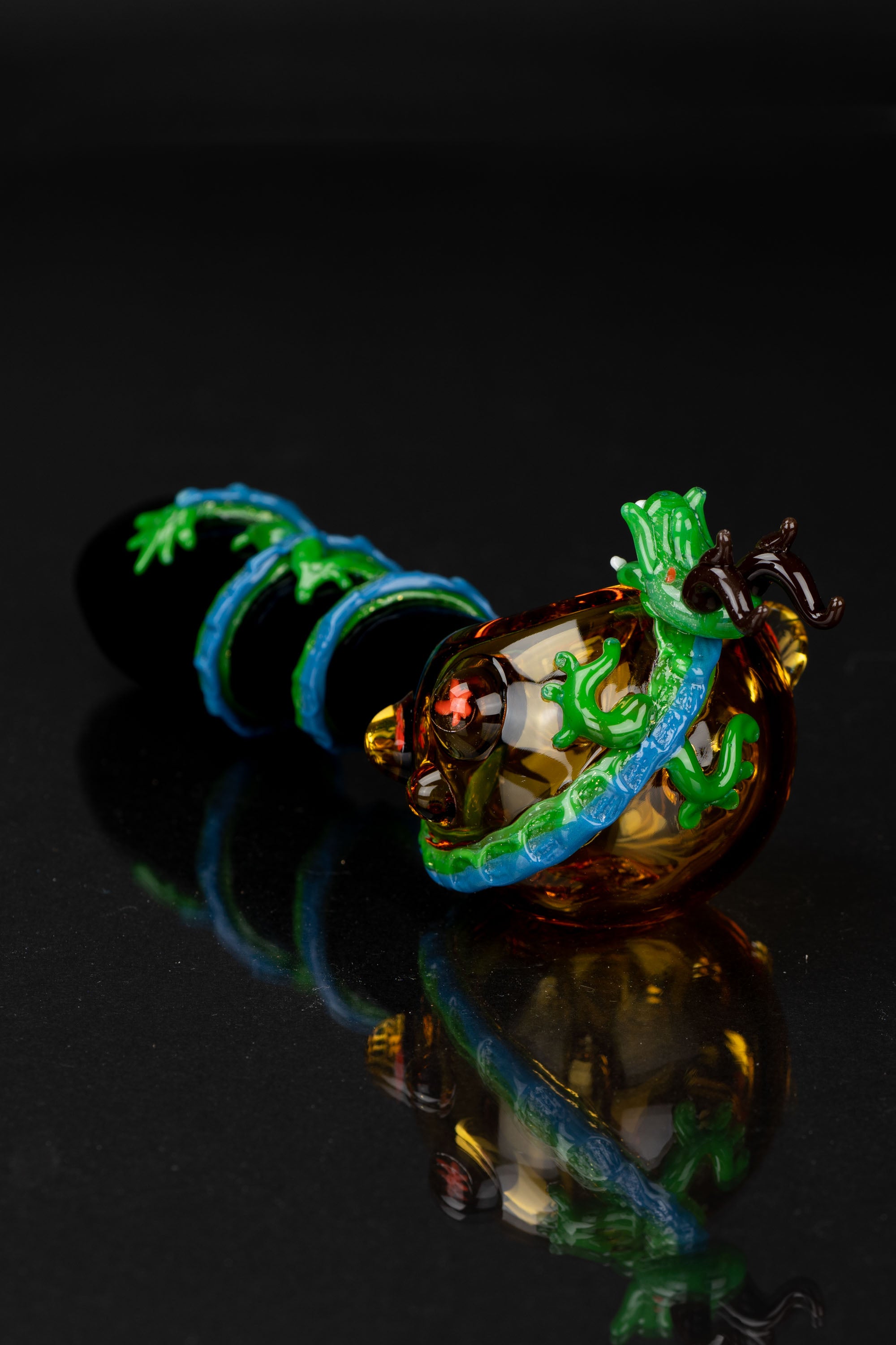 Hand-blown glass cannabis pipe with intricate amber, turquoise, and green floral detailing, displayed on a reflective black surface.