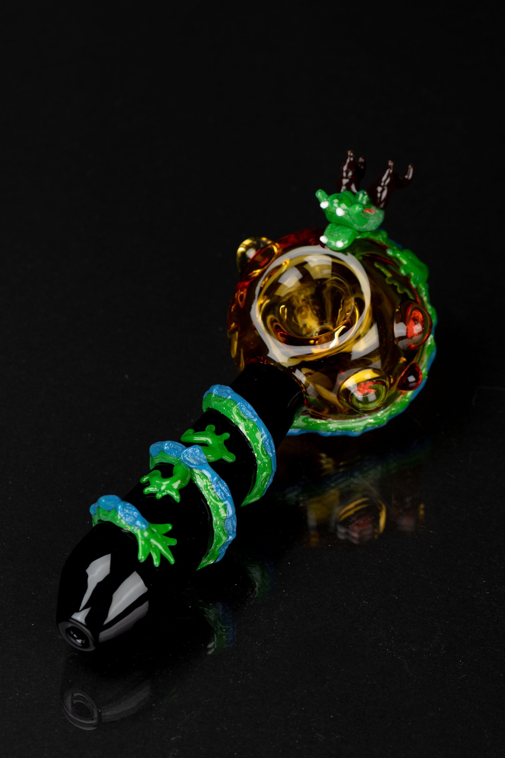 Hand-blown glass pipe with a vibrant green lizard design and intricate detailing, displayed on a reflective black surface.