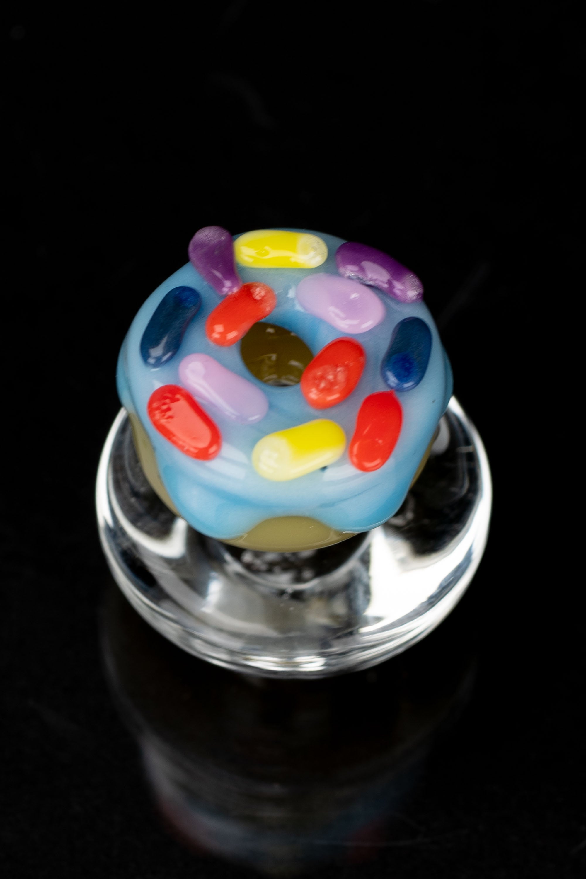 A colorful glass cannabis oil rig topper with scattered, jewel-toned glass “drops” against a black background.