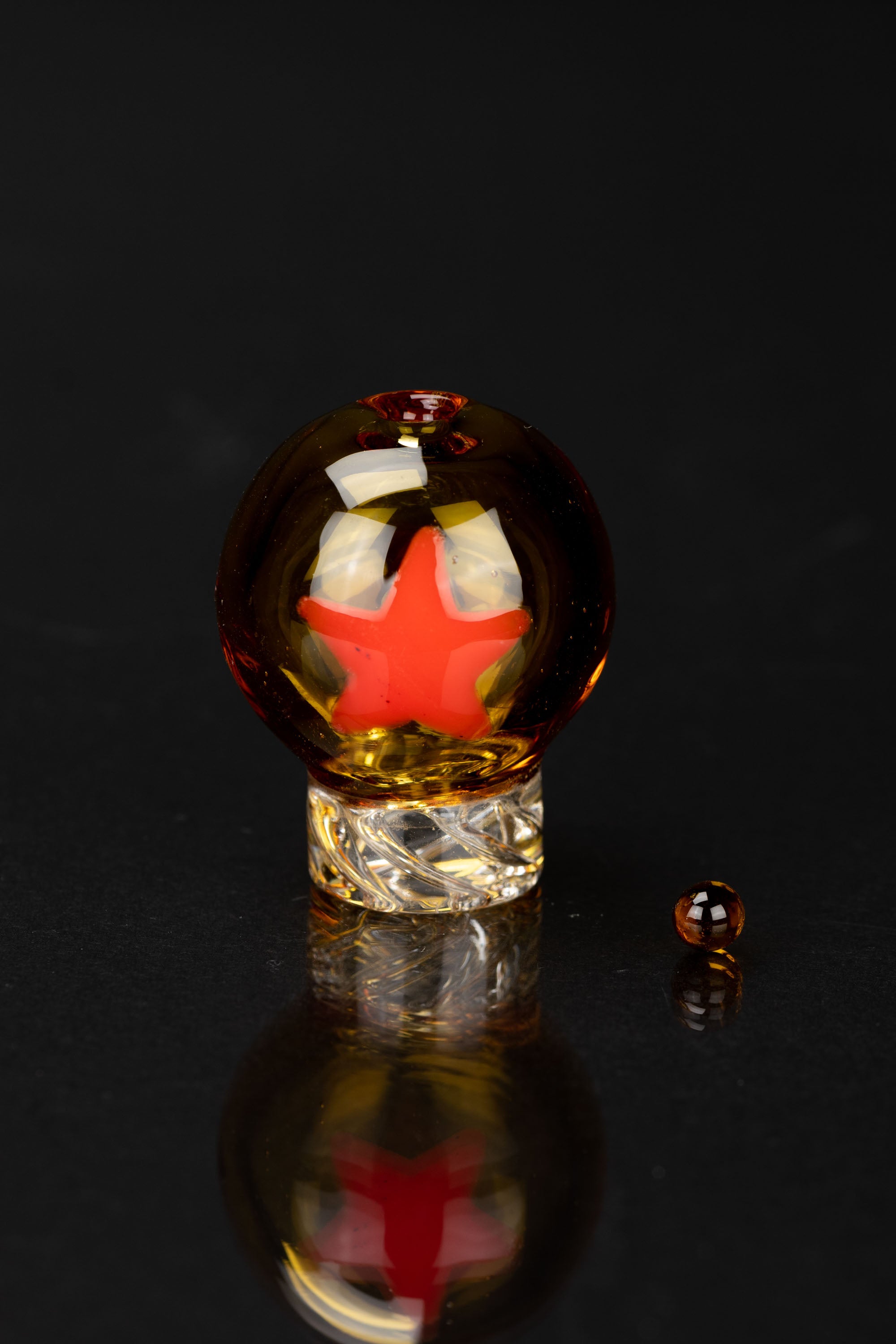 A glass dome with a red starburst pattern inside, resting on a textured base, with a small glass bead beside it, all reflected on a dark surface.