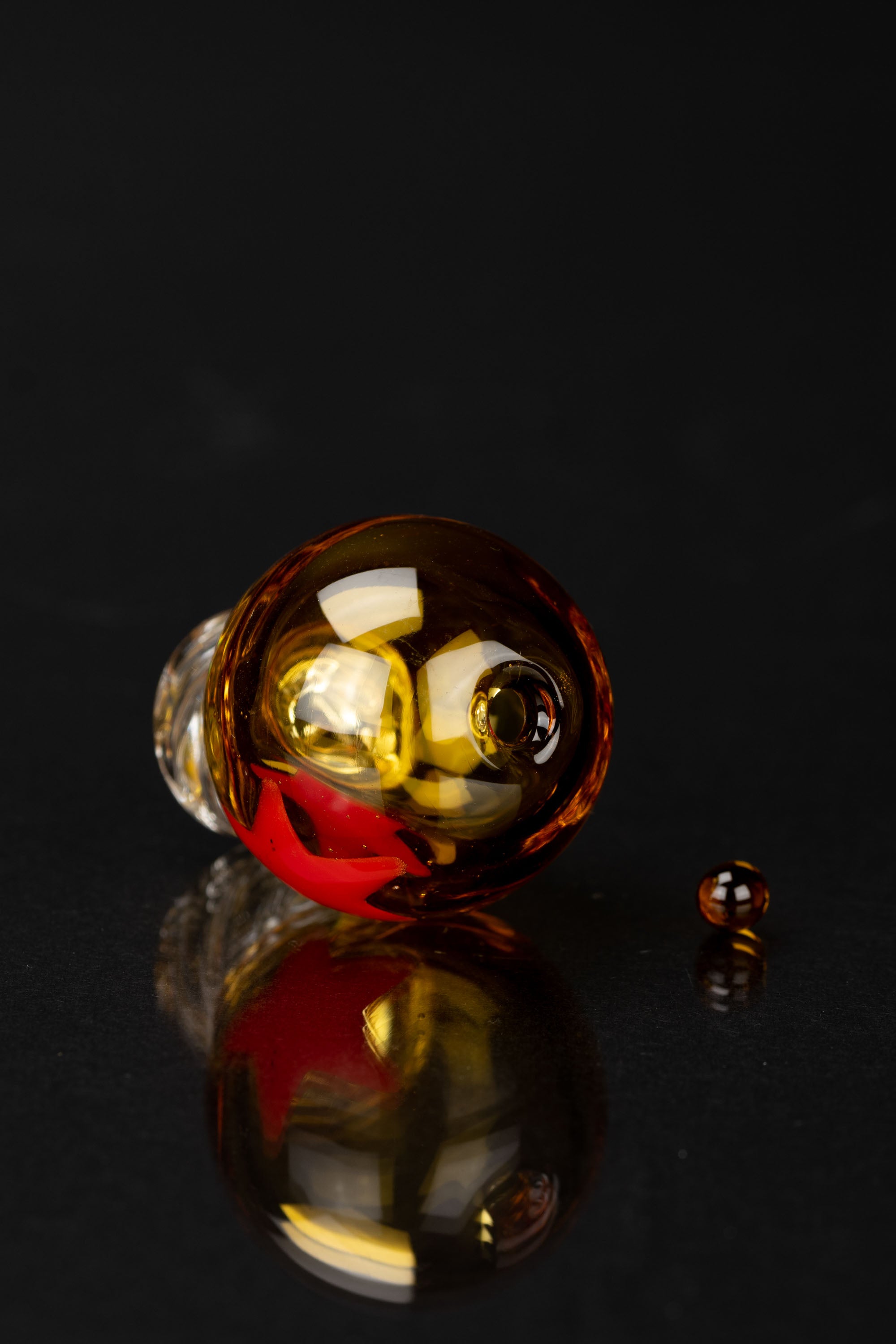 A vibrant glass cannabis water pipe with red and amber hues, reflected on a dark surface.