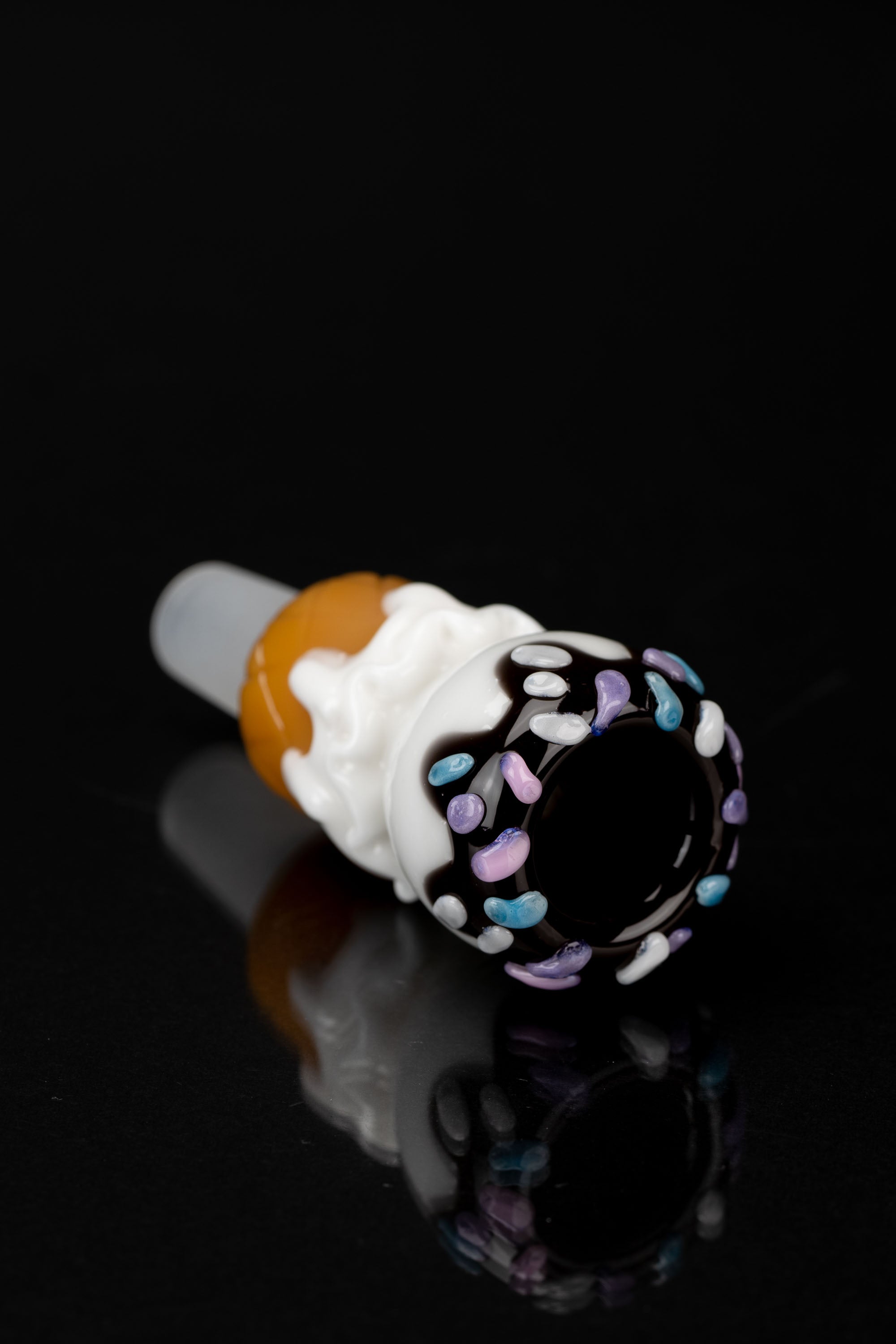 Hand-blown glass cannabis pipe resembling a melting ice cream cone with colorful sprinkles, against a black reflective surface.