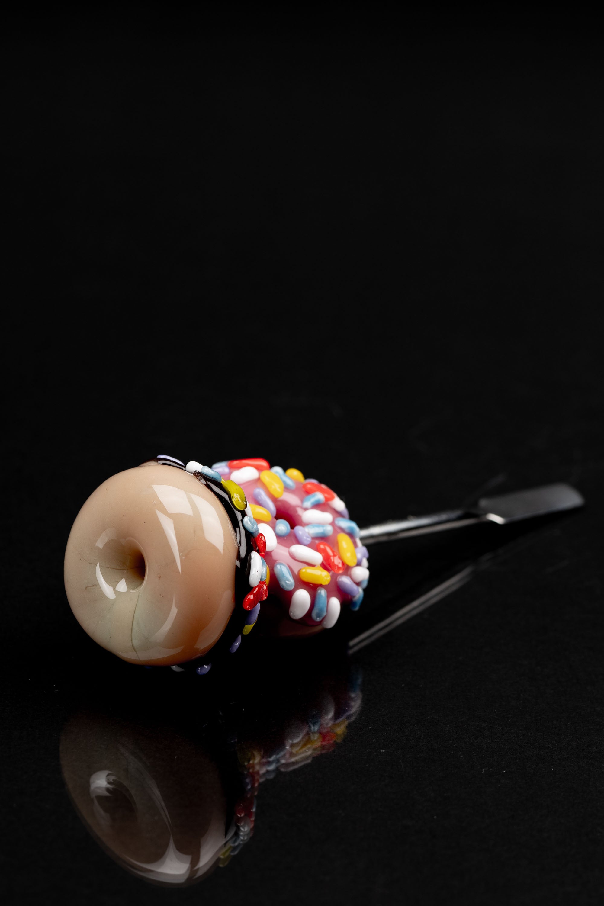 Glass pipe shaped like a frosted donut with colorful sprinkles, reflecting on a black surface.