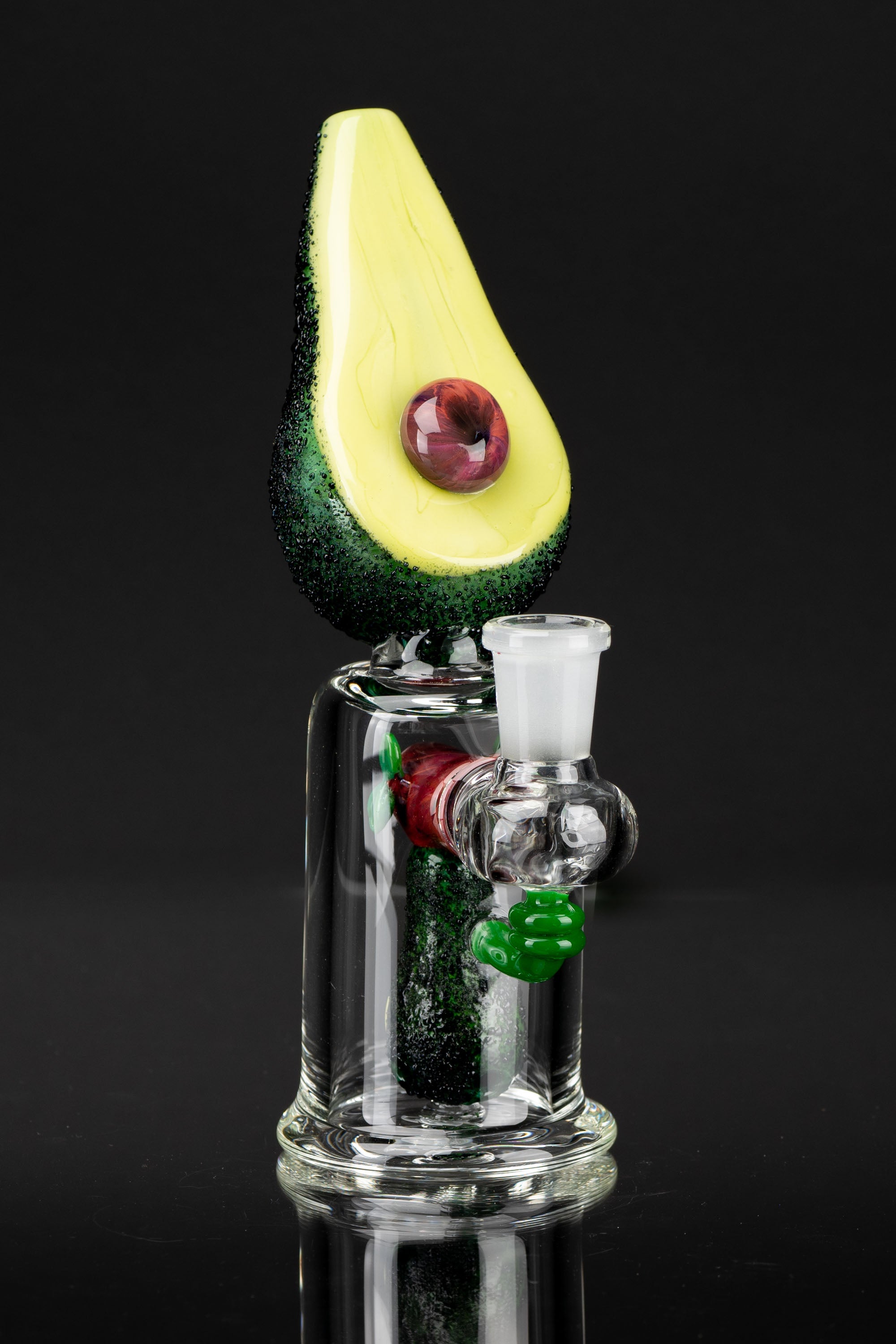 A glass water pipe shaped like an avocado with a red “pit” and green accents, against a black background.