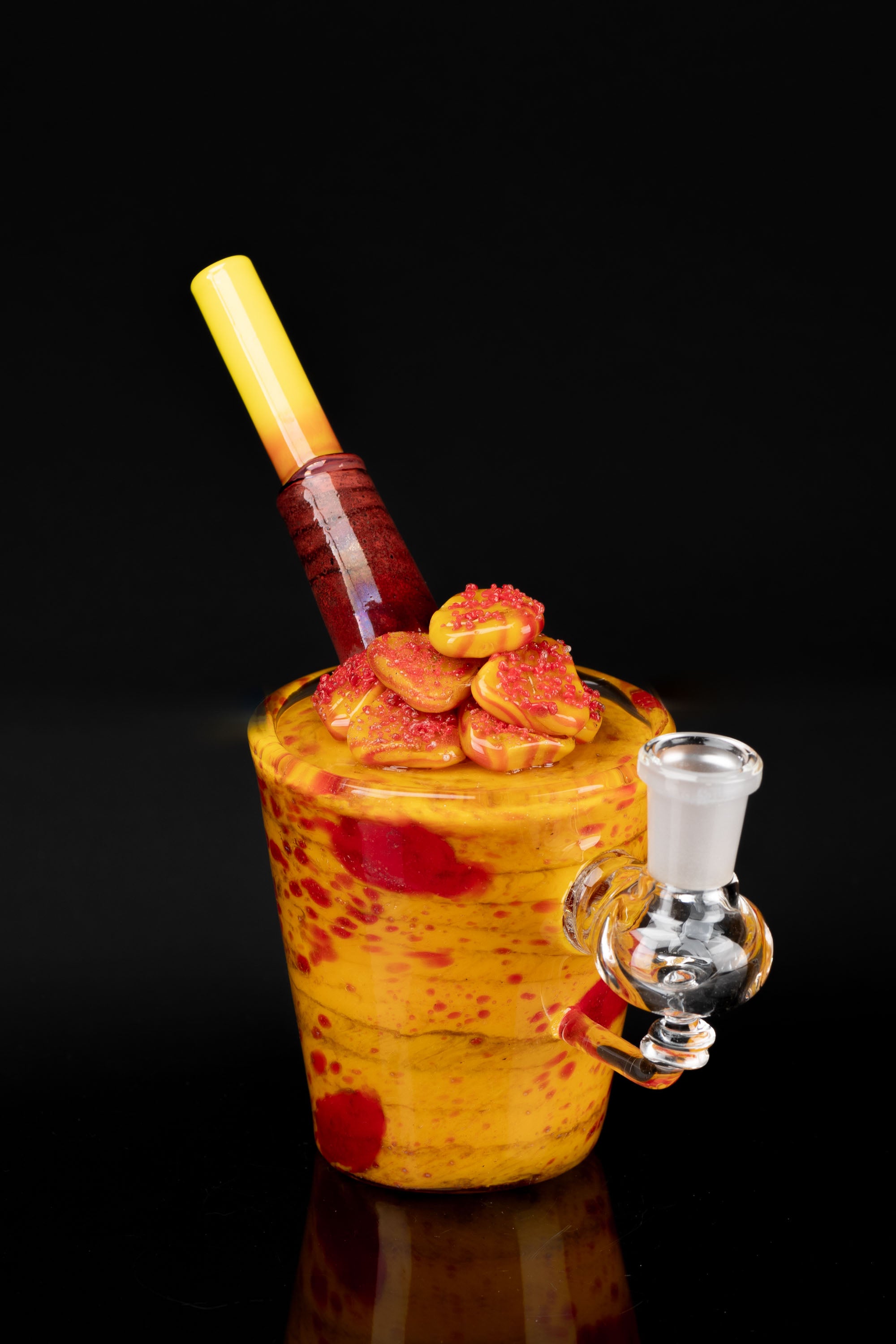 A vibrant, hand-blown glass water pipe resembling a dessert with layered orange and red hues, topped with sculpted glass candies, and featuring a clear glass mouthpiece, set against a black background.