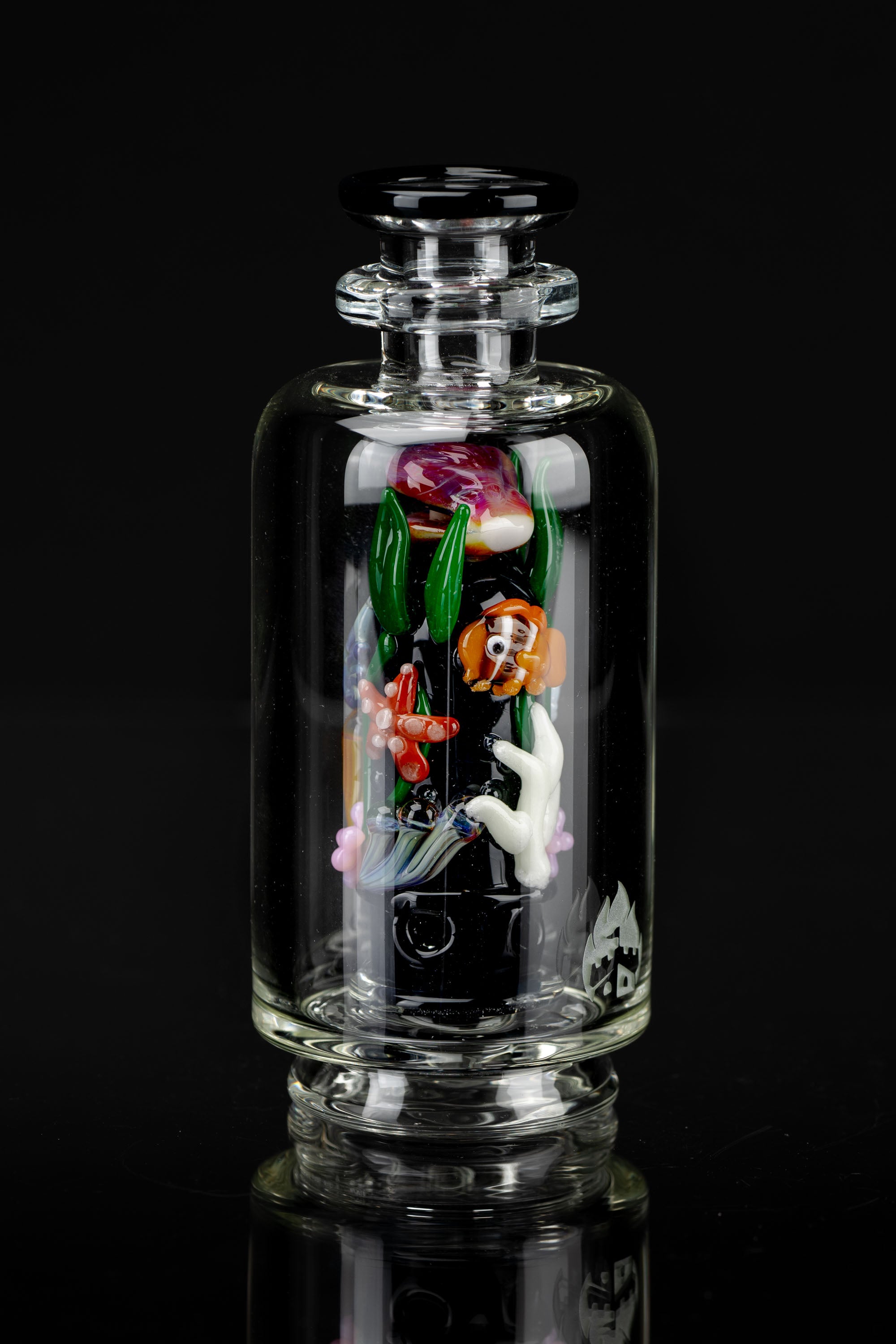 A clear glass bubbler with colorful sculpted glass figures of koi fish and floral elements inside, reflected on a glossy black surface.