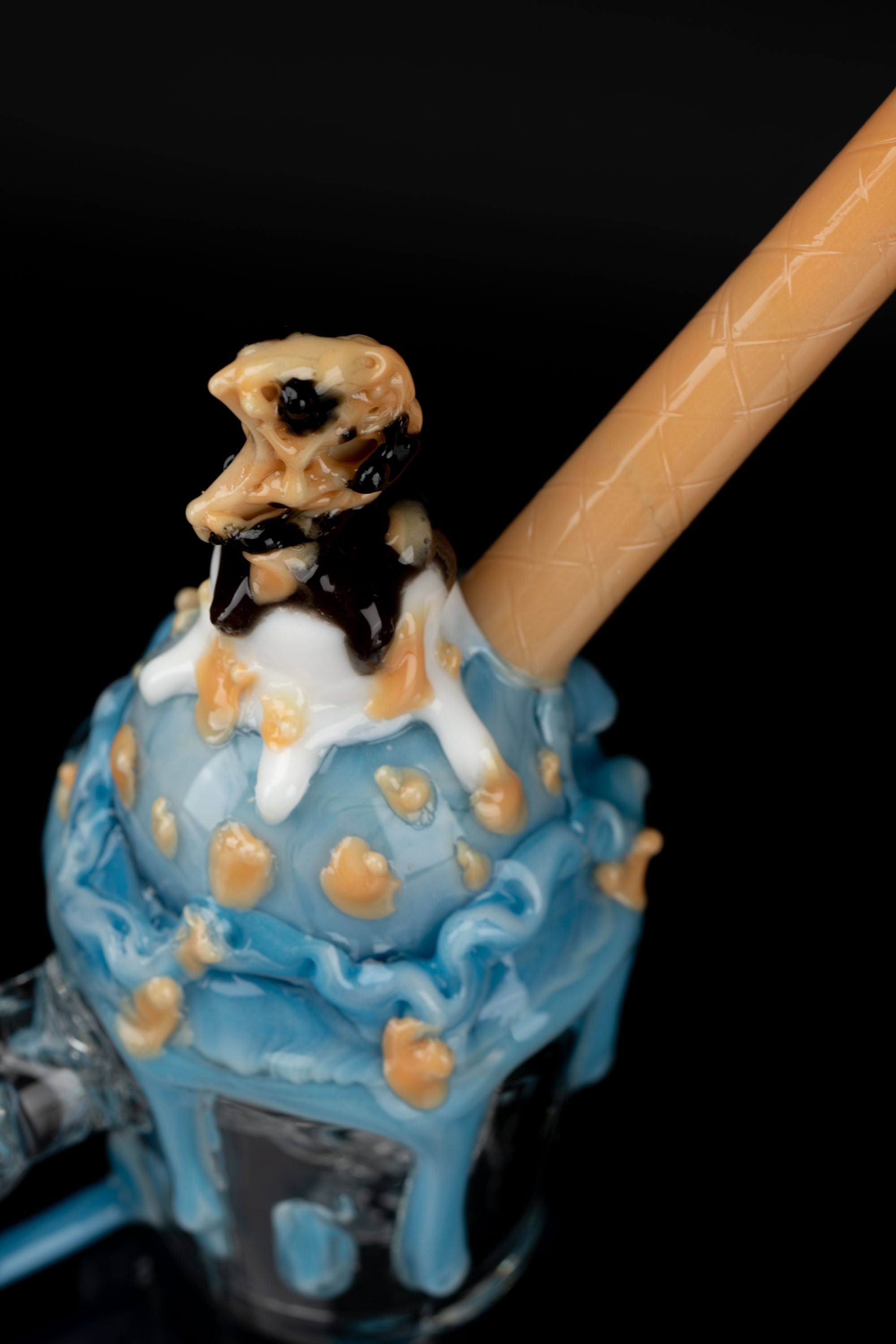 A detailed glass cannabis water pipe resembling a dripping ice cream sundae with chocolate and caramel accents, against a black background.