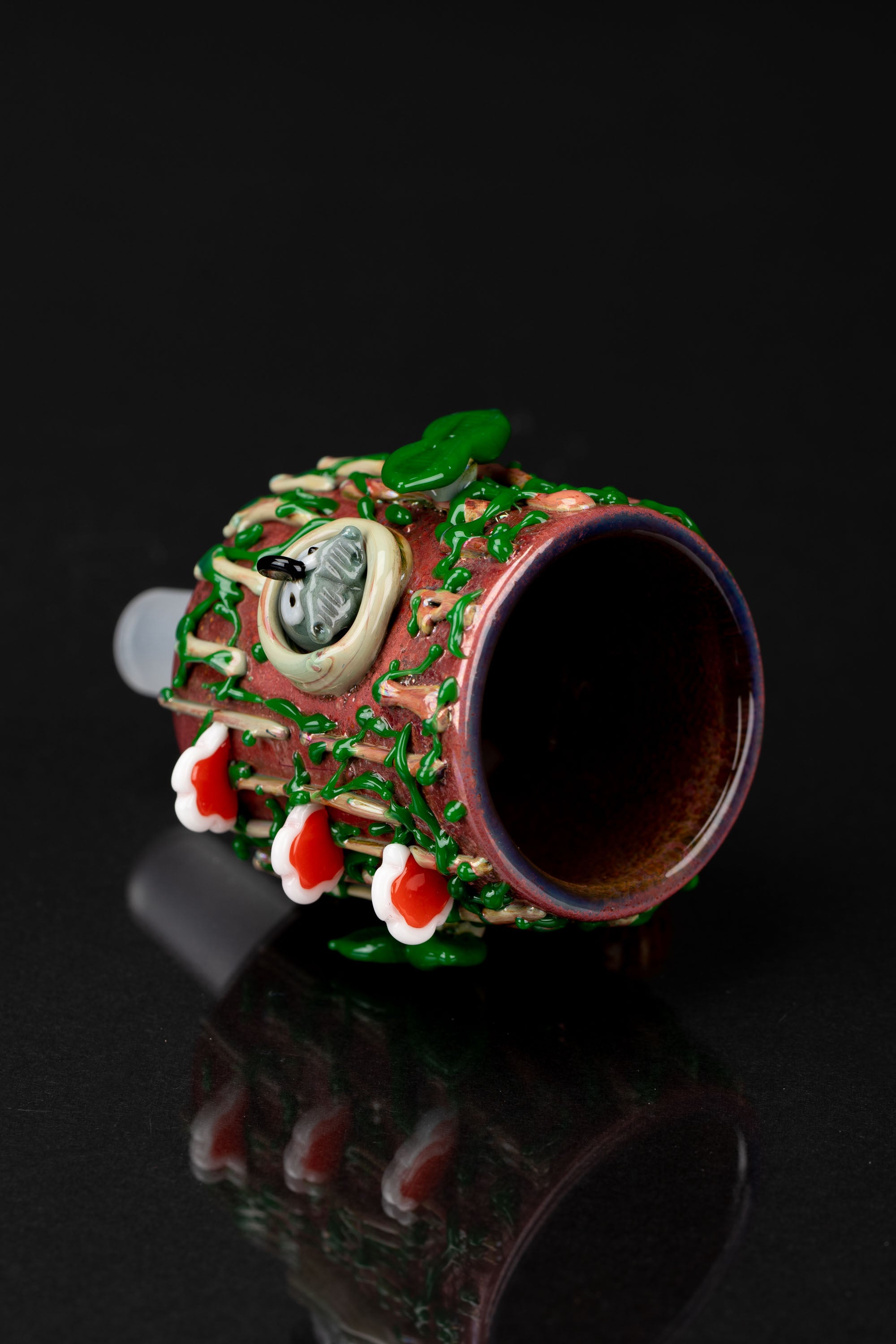 Hand-blown glass pipe with a sculpted, vibrant green and red design, resembling a fantastical creature or plant, against a black background.