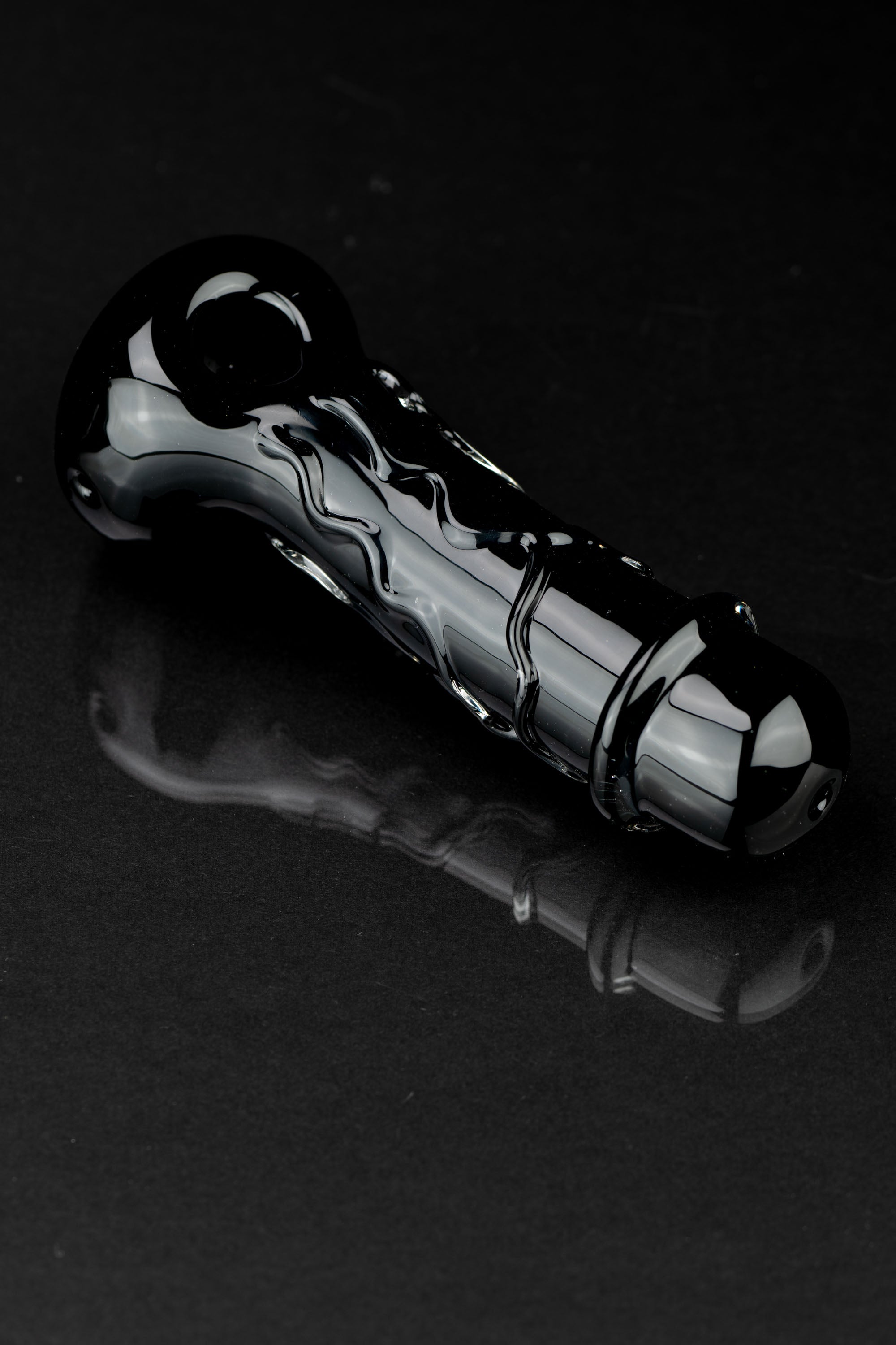 Black glass cannabis pipe with swirling design, reflecting on a dark surface.