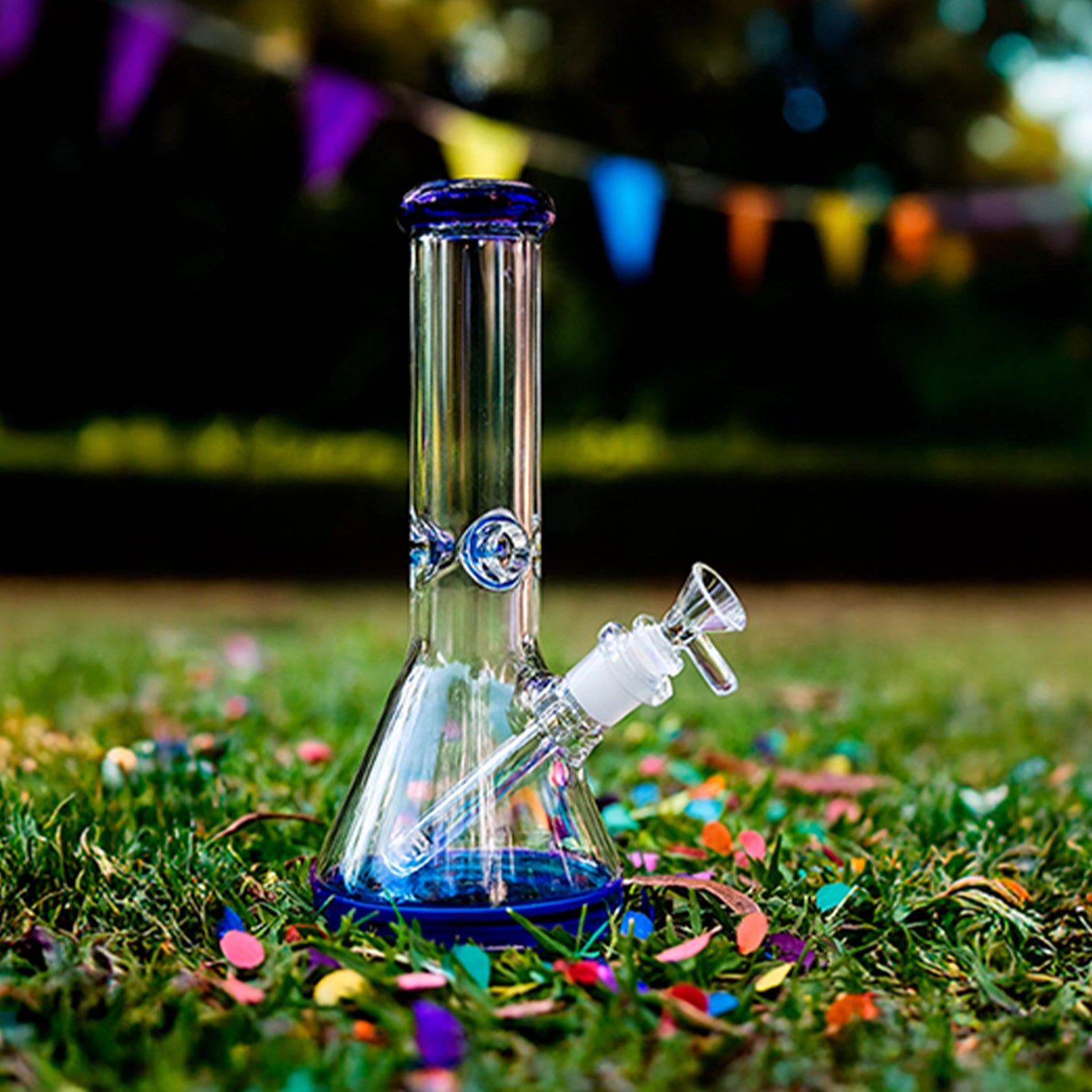 Front view of the Smoke Cartel 9mm Big Base Beaker w/ Color Accents Bong - 12 in. resting on grass with blue-accented glass and colorful confetti.