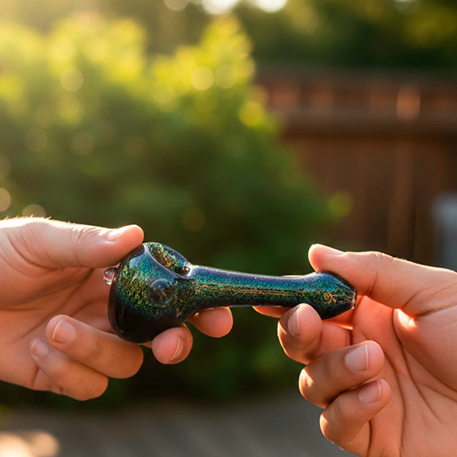 Close-up of two hands passing the LA Pipes Solid Blue Dichro Spoon - Blue Dream, showing its iridescent blue-green finish and rounded bowl against a sunlit blurred garden background