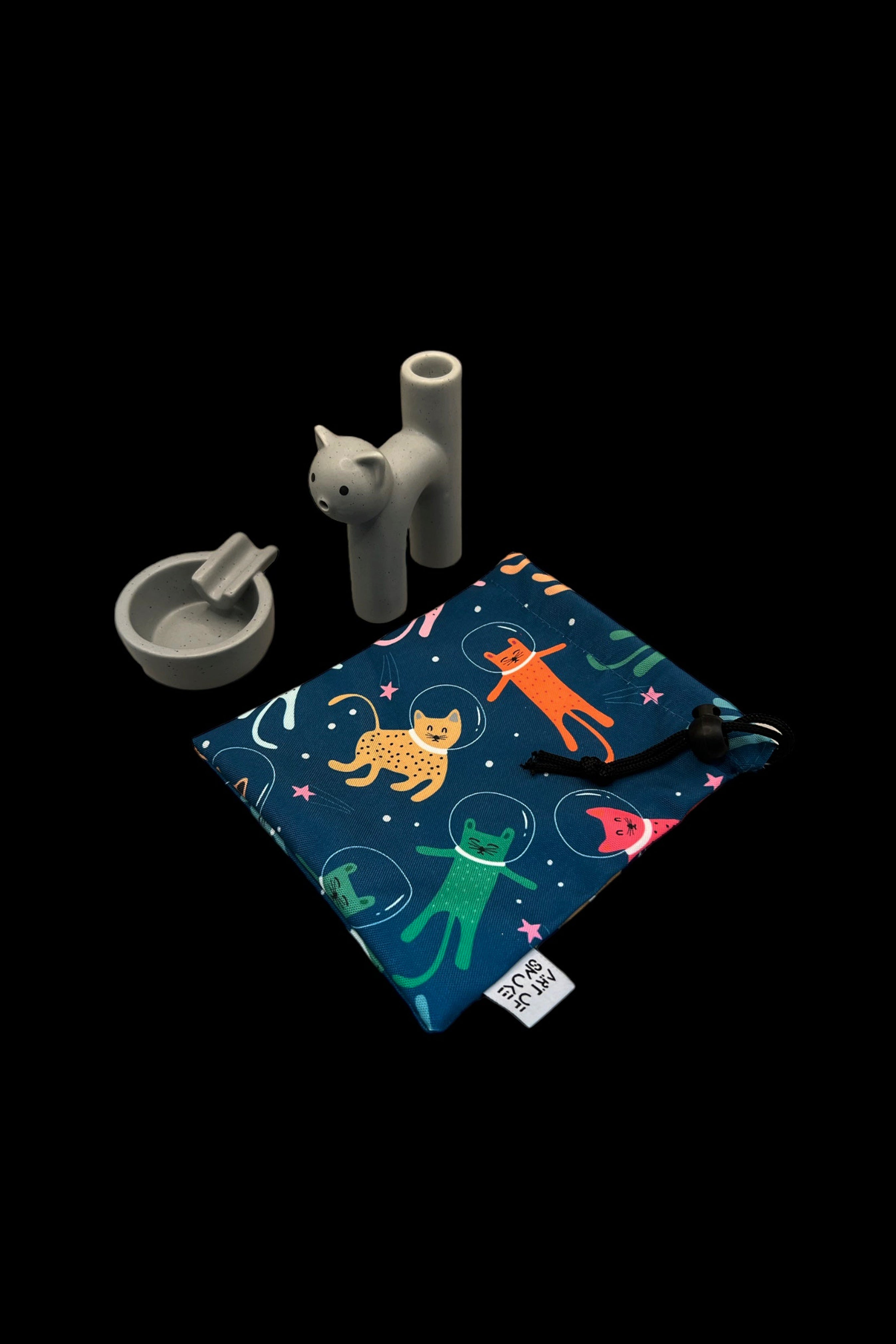 A colorful, patterned cannabis accessories pouch with a space cat design, displayed with a grinder and pipe on a dark background.