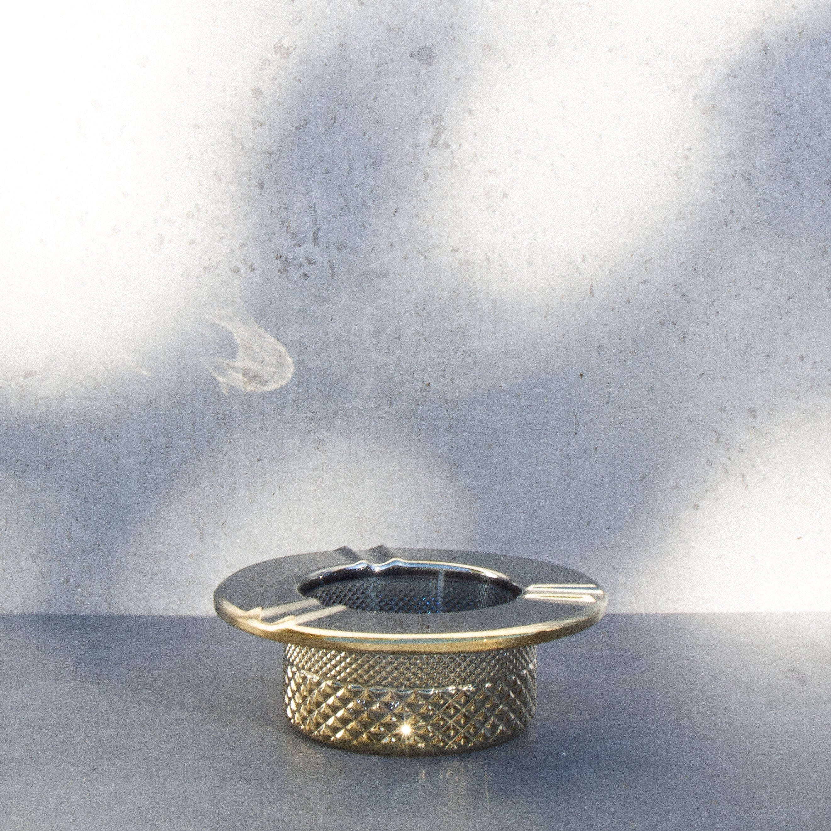 Gold cannabis ashtray with a textured, patterned base, set against a muted, textured wall.