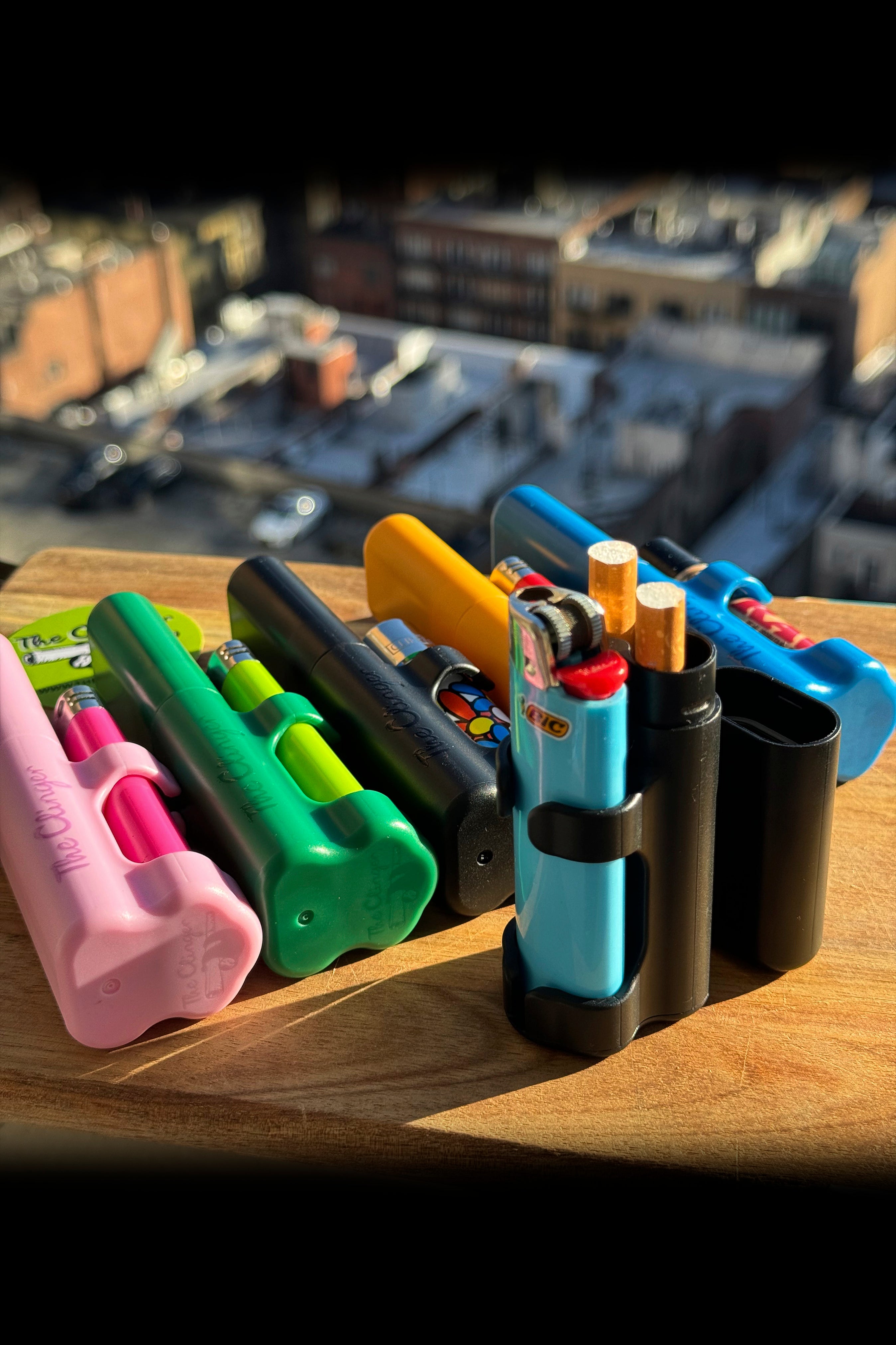 Colorful silicone sleeves protecting cannabis vapes and lighters on a wooden surface, with a blurred cannabis display in the background.