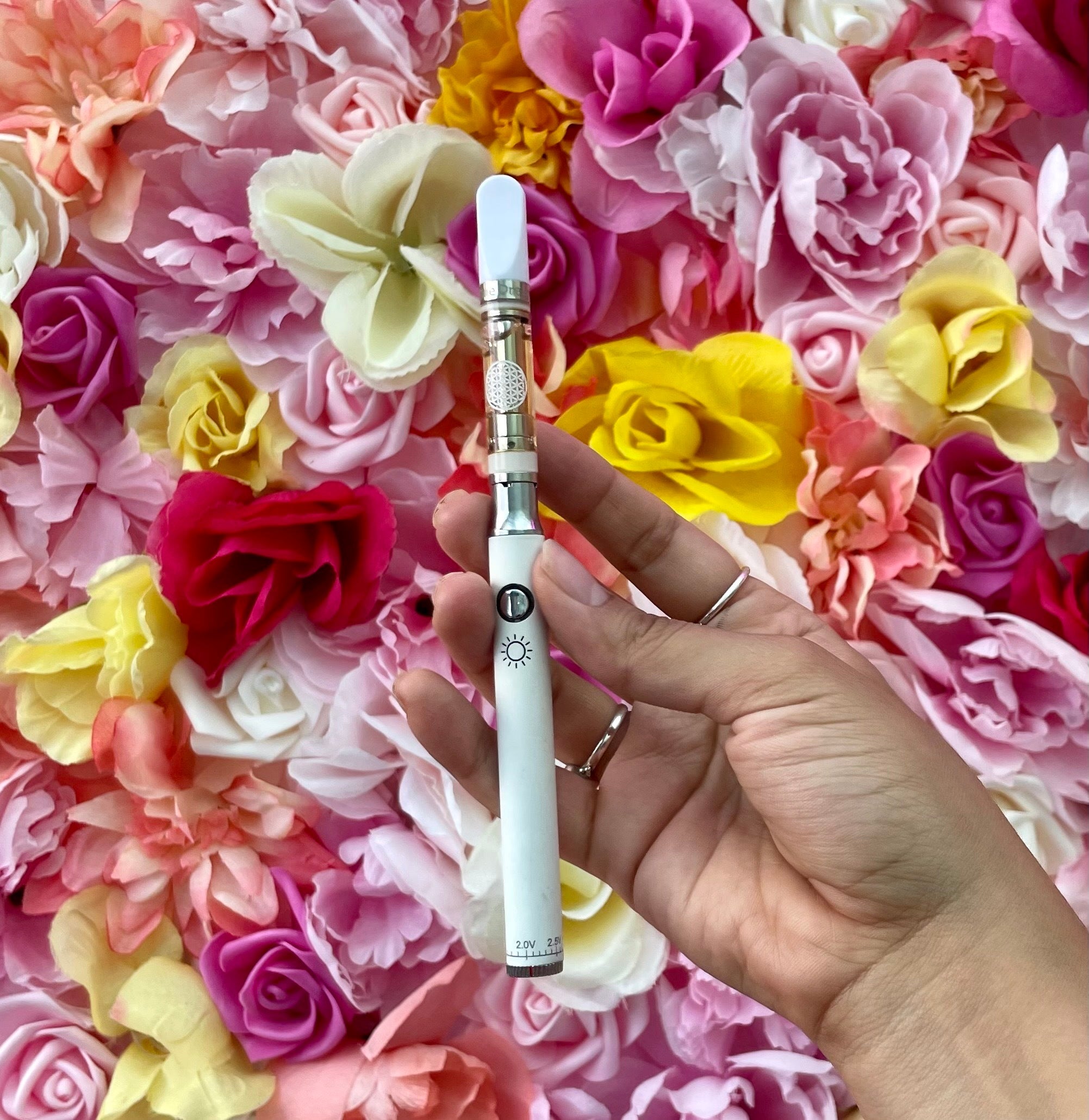 White cannabis vape pen resting on a bed of pink and yellow roses. <start_of_image>