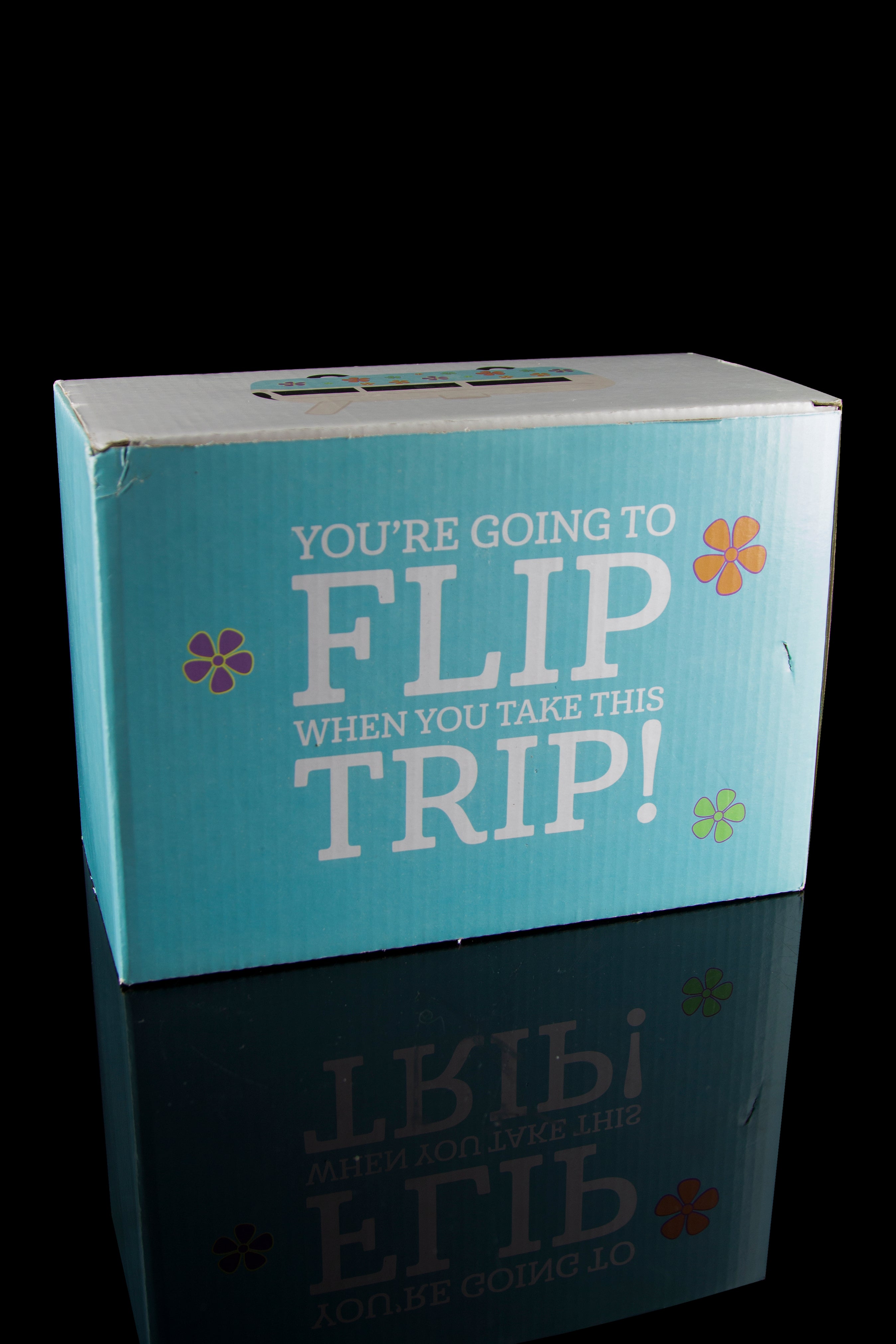 A teal cannabis accessory box with floral accents reads "You're Going to Flip When You Take This Trip!" reflected on a glossy surface.
