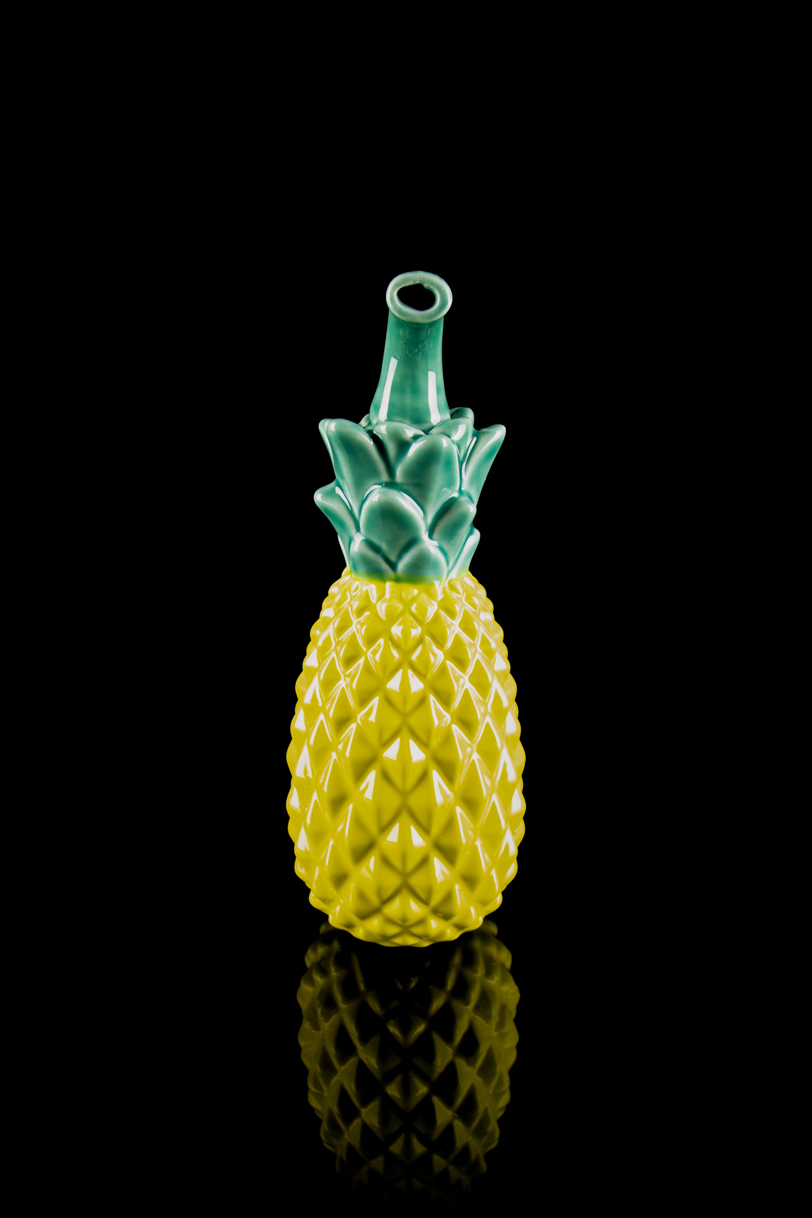 Yellow glass pineapple-shaped water pipe with a green leafy top, reflecting on a black surface.