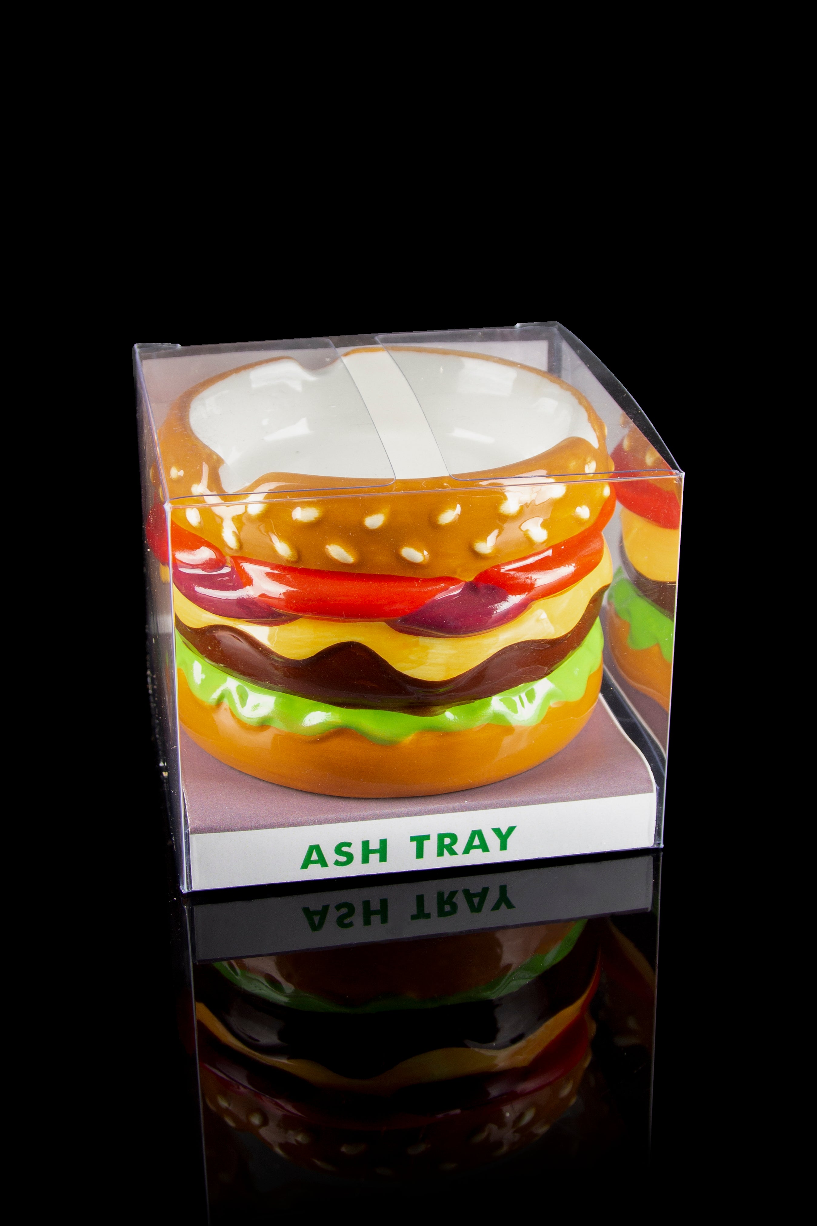 A clear acrylic ashtray shaped like a colorful hamburger with a green “ASH TRAY” label, reflected on a black surface.