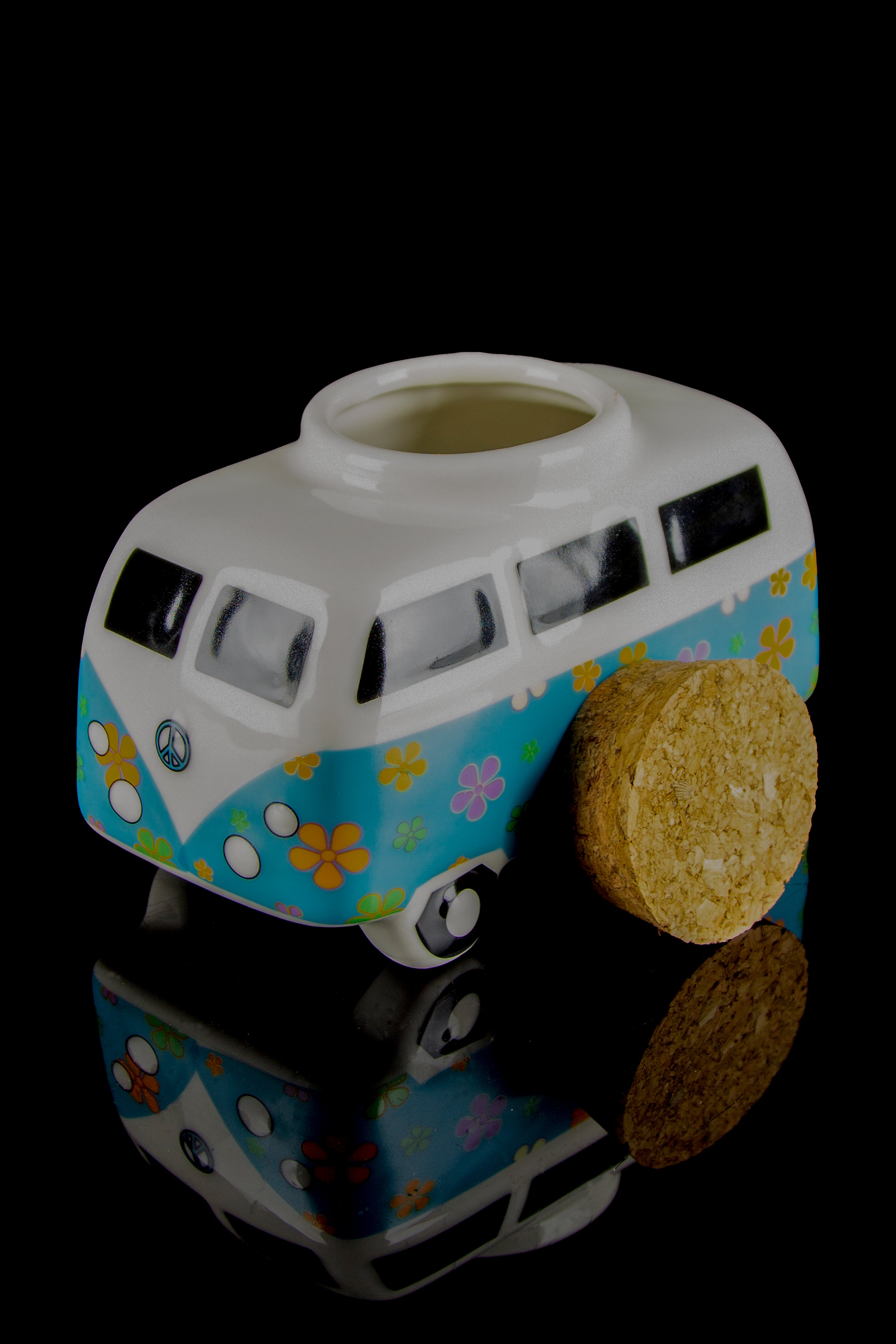 A colorful, ceramic cannabis water pipe shaped like a vintage VW bus with two spherical hash balls resting against it, all reflected on a black surface.