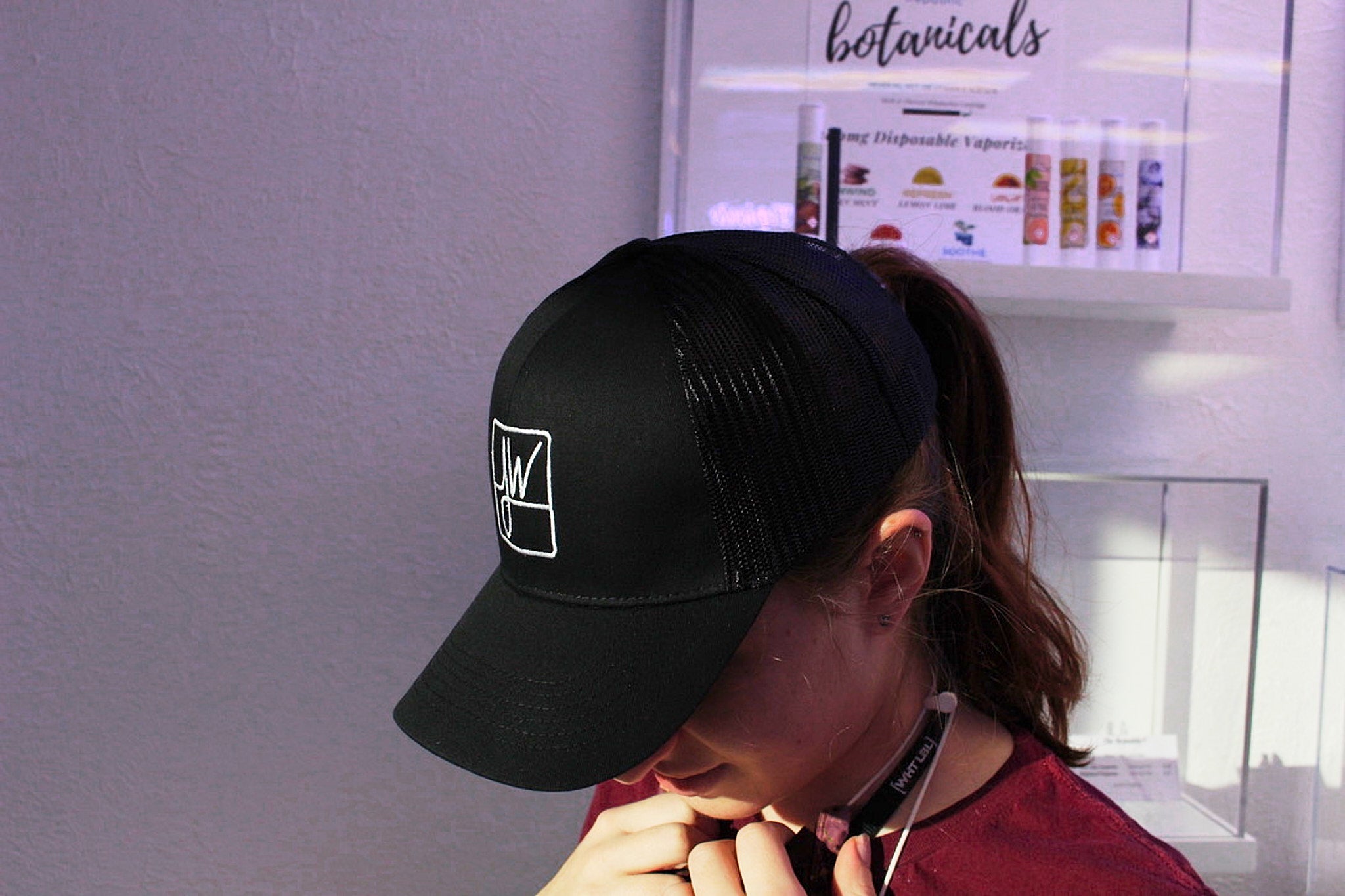 A person wearing a black trucker hat with a white logo, standing in front of a display of cannabis vaporizers.