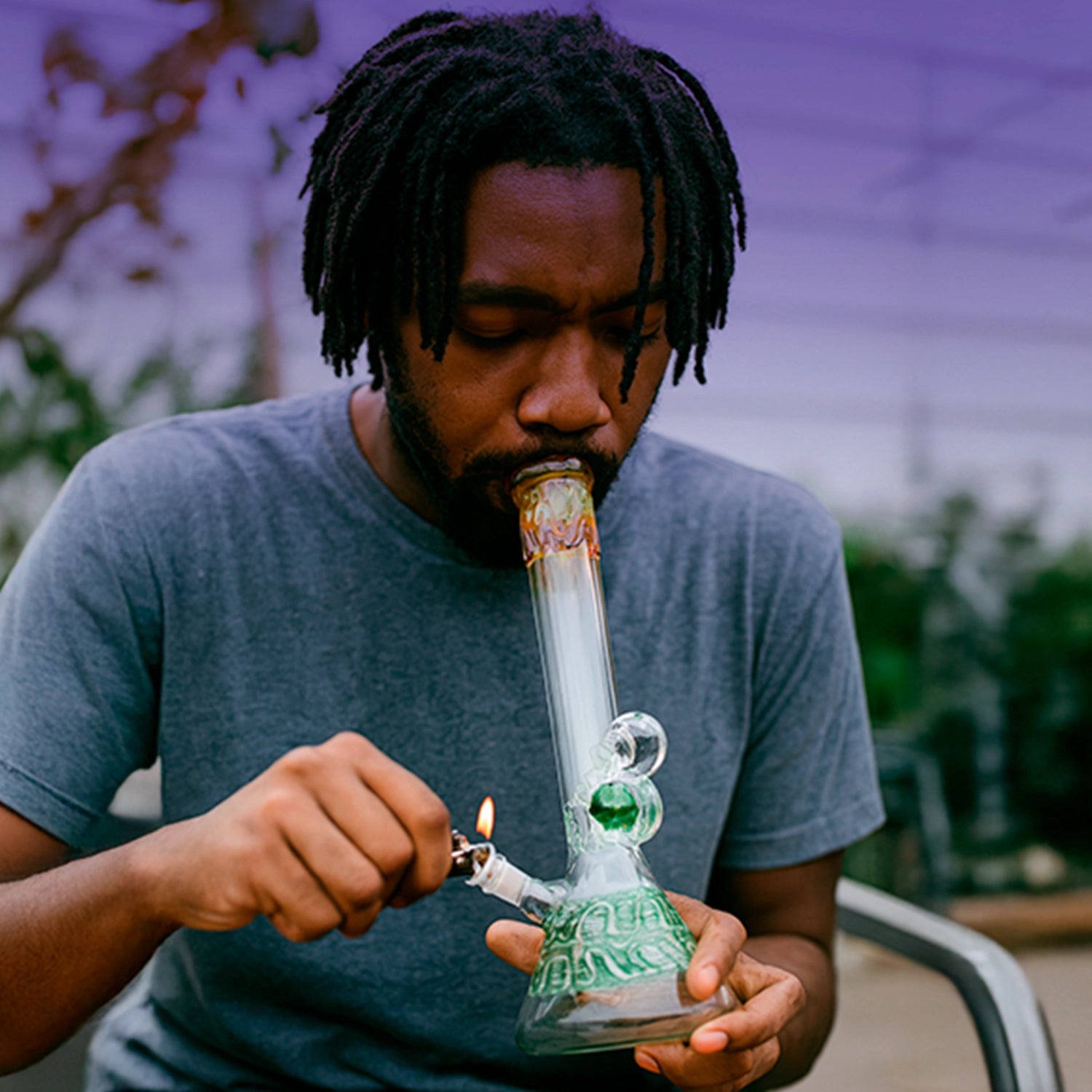 Close-up of a man lighting the mouthpiece of a Smoke Cartel Mushroom Marble Water Pipe - 18 in., shown from a frontal angle against a blurred backyard background.