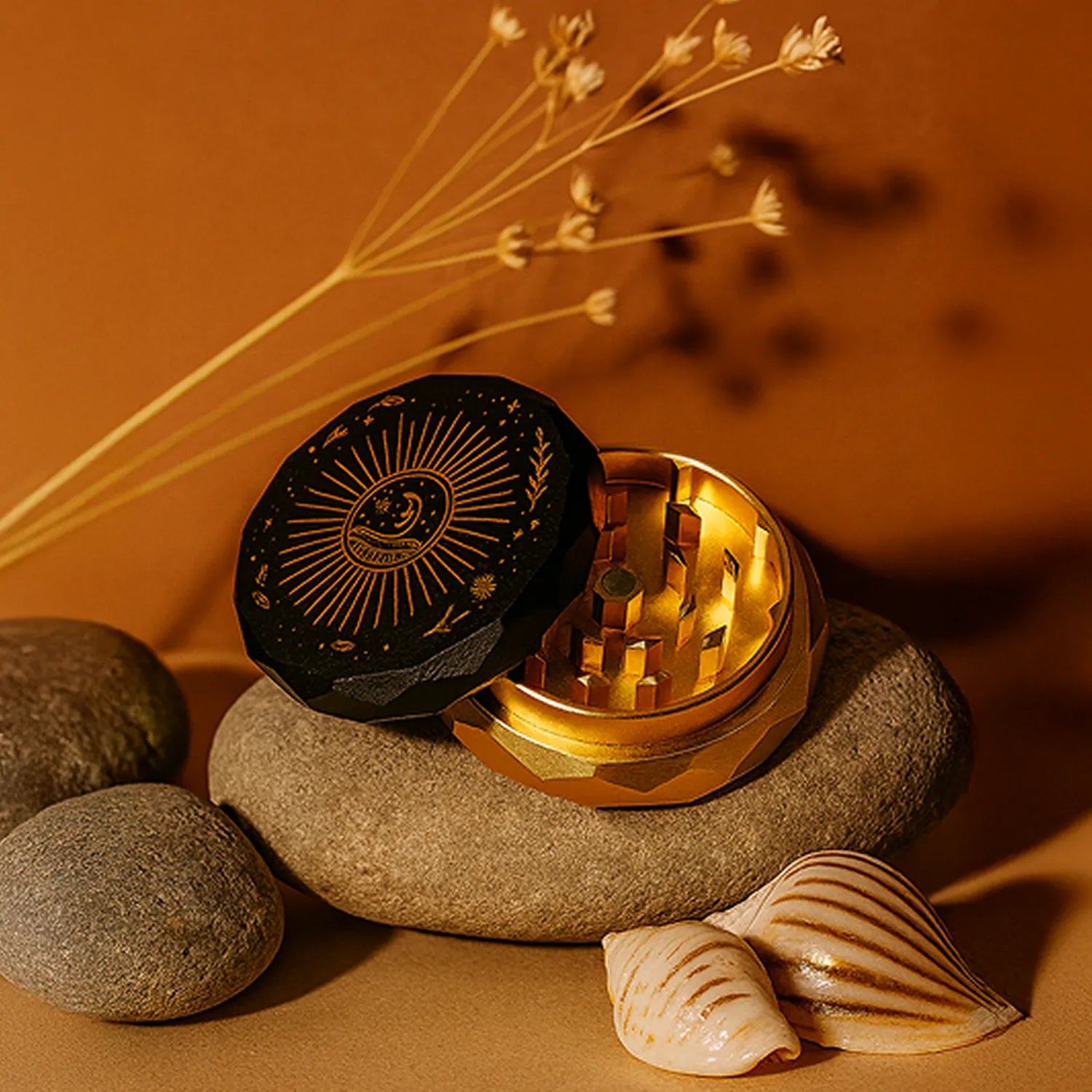 Diagonal close-up of the black and gold Famous Design - Astral Power Aluminum 2 stage Grinder - Black and Gold resting open on smooth stones with seashells and dried stems.