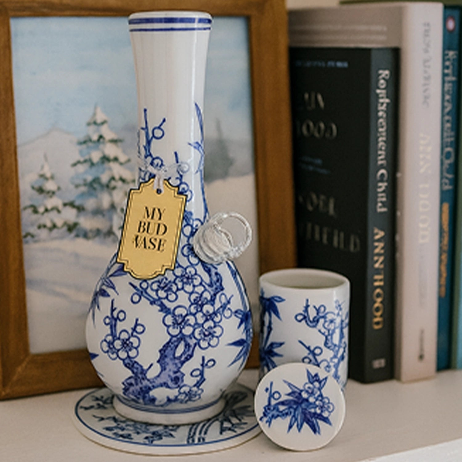 Close-up front view of the My Bud Vase "Luck" Porcelain Vase Bong adorned with blue floral branches and a hanging gold tag, set against books and a framed winter print