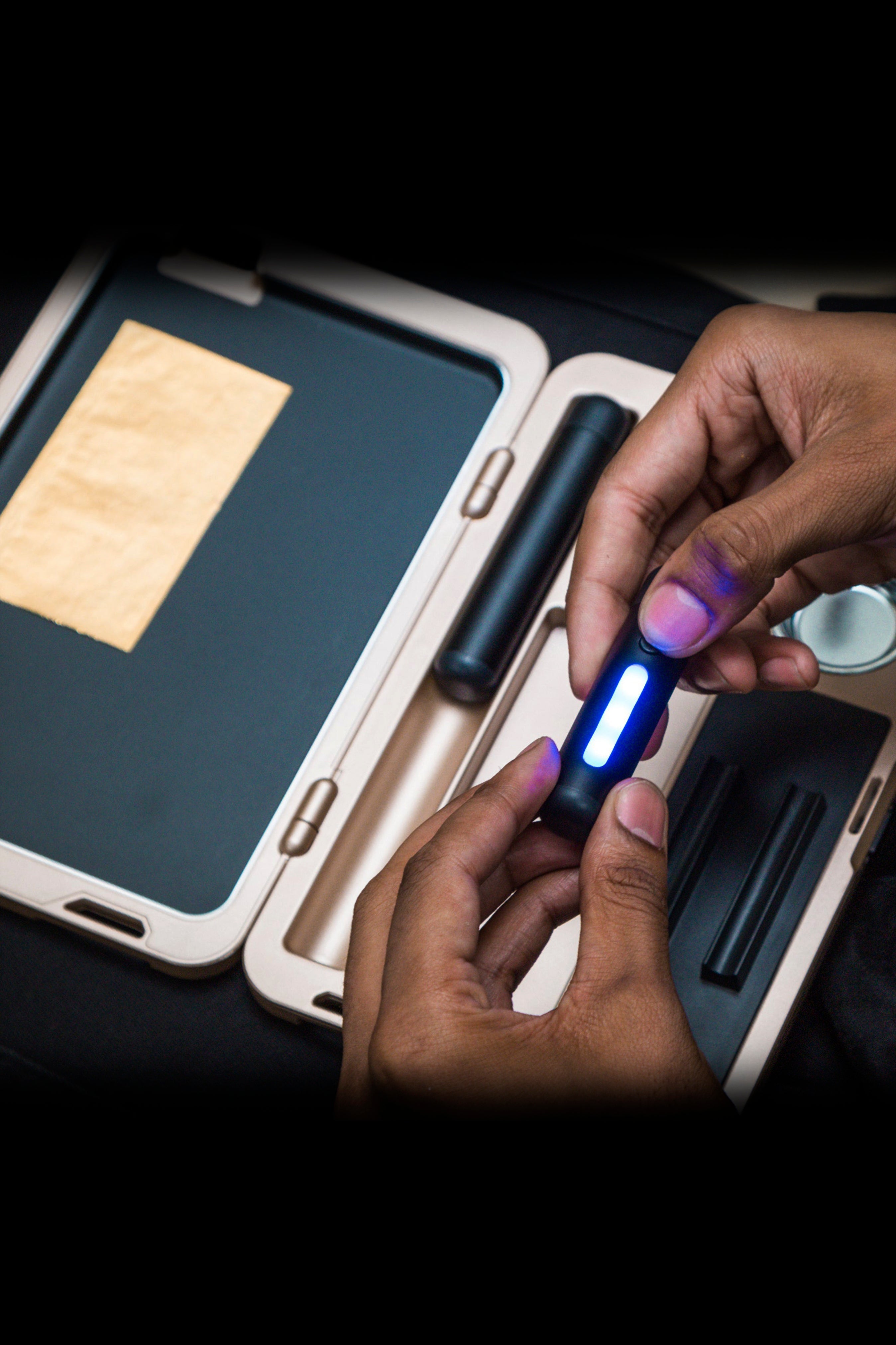Hands holding a lit, glowing cannabis vaporizer pen over an open, wood-toned cannabis accessory case with rolled joints and a grinder inside.