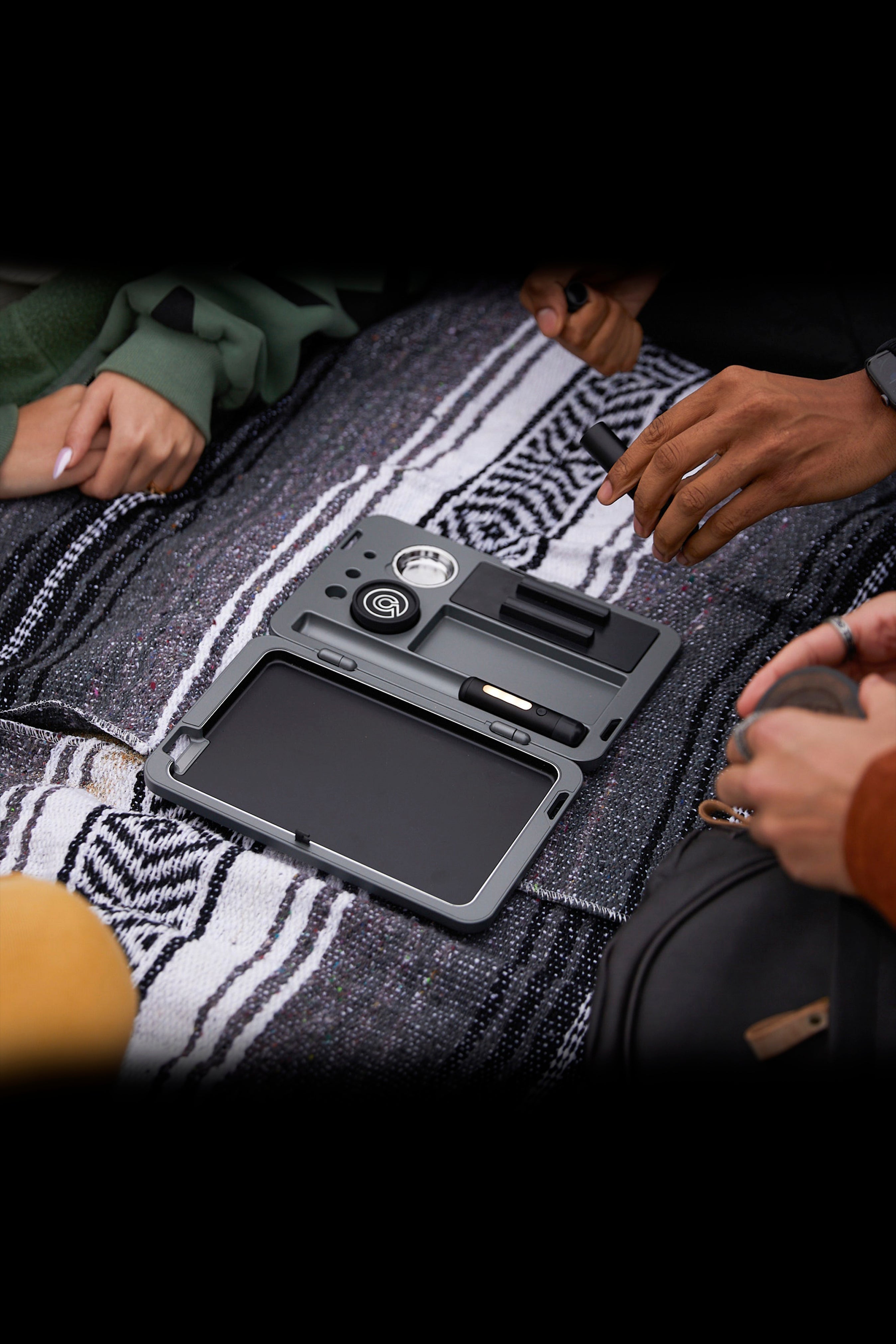 Overhead view of a cannabis storage case open on a patterned blanket, surrounded by people's hands and clothing.