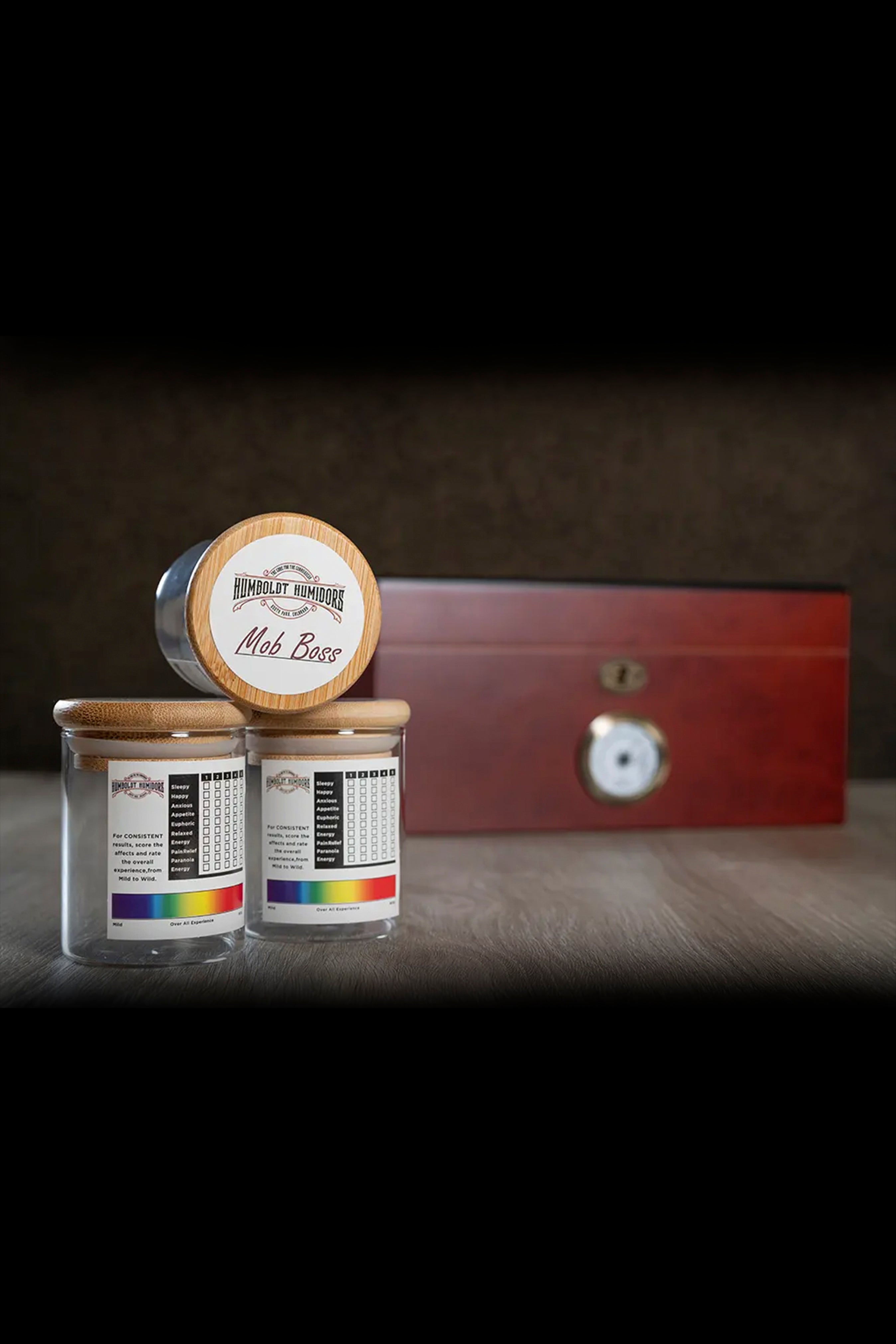 A still life of cannabis accessories including a wooden humidor labeled “Mob Boss”, two glass jars with colorful cannabis strain information, and a small metallic sculpture, all on a reflective black surface.