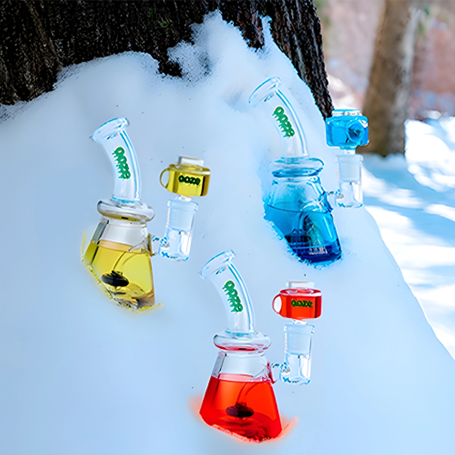 Tilted close-up of colorful glass pieces of the Ooze Glyco Glycerin Chilled Bong set embedded in snow against a tree trunk background