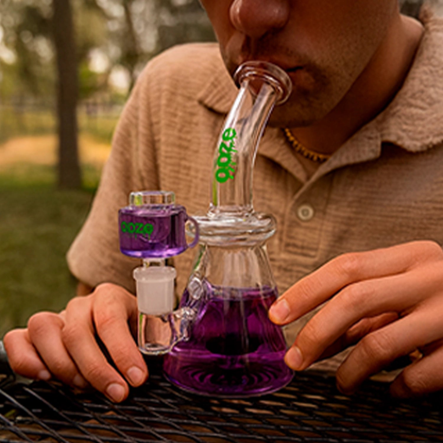Close-up of a person using an Ooze Glyco Glycerin Chilled Bong, angled mouthpiece and purple water visible against a blurred outdoor background