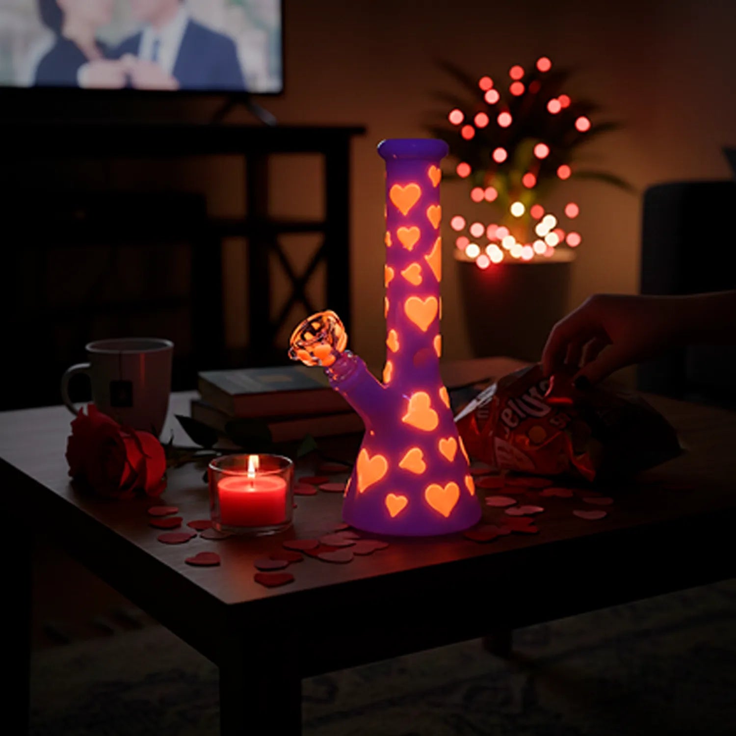 A dimly lit front view of the Valentines Day Heart Glow In The Dark Beaker glowing purple with orange heart cutouts on a coffee table amid rose petals and a lit candle