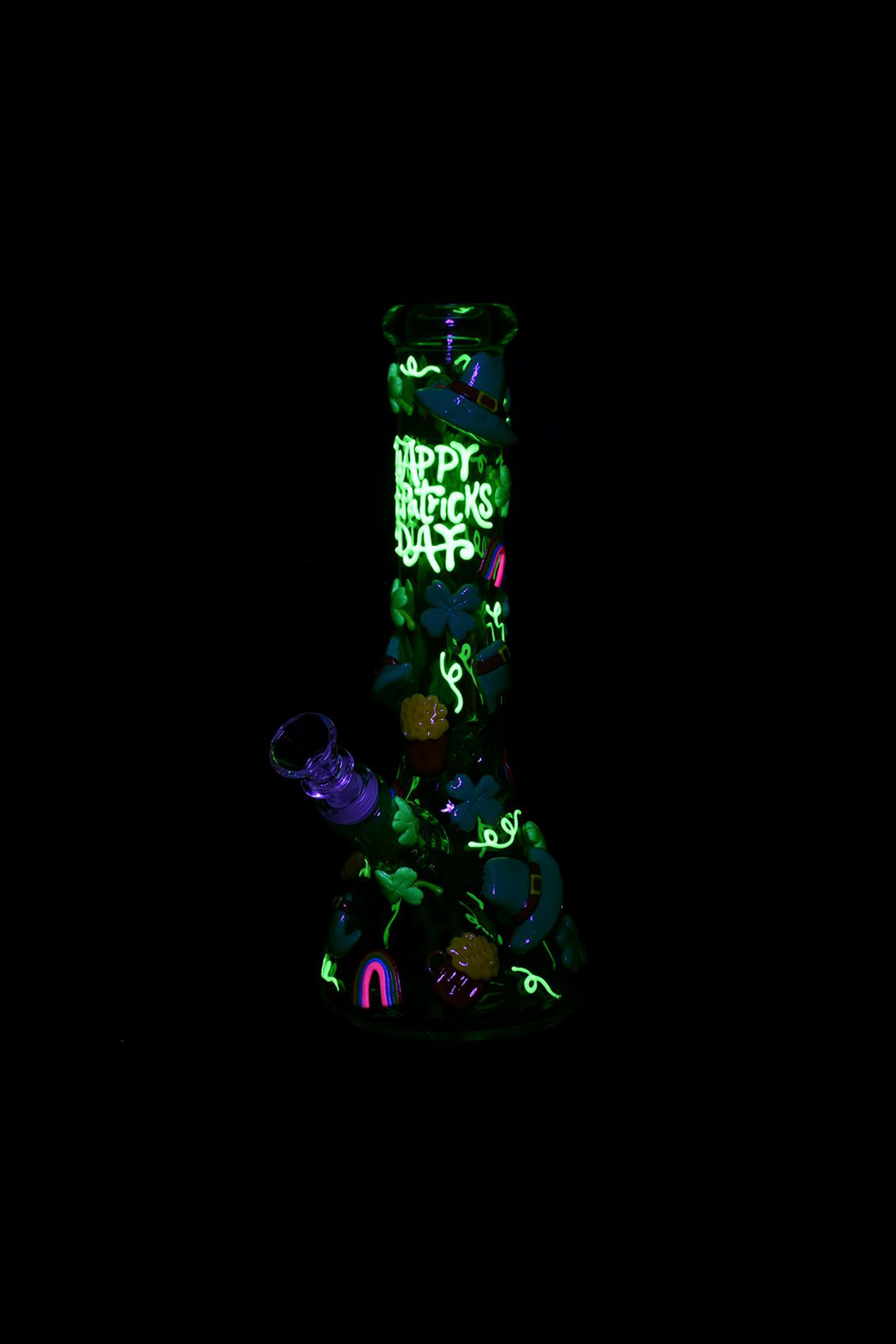 A black glass cannabis water pipe decorated with green shamrocks and "Happy St. Patrick's Day" lettering, illuminated with neon green and purple lights.