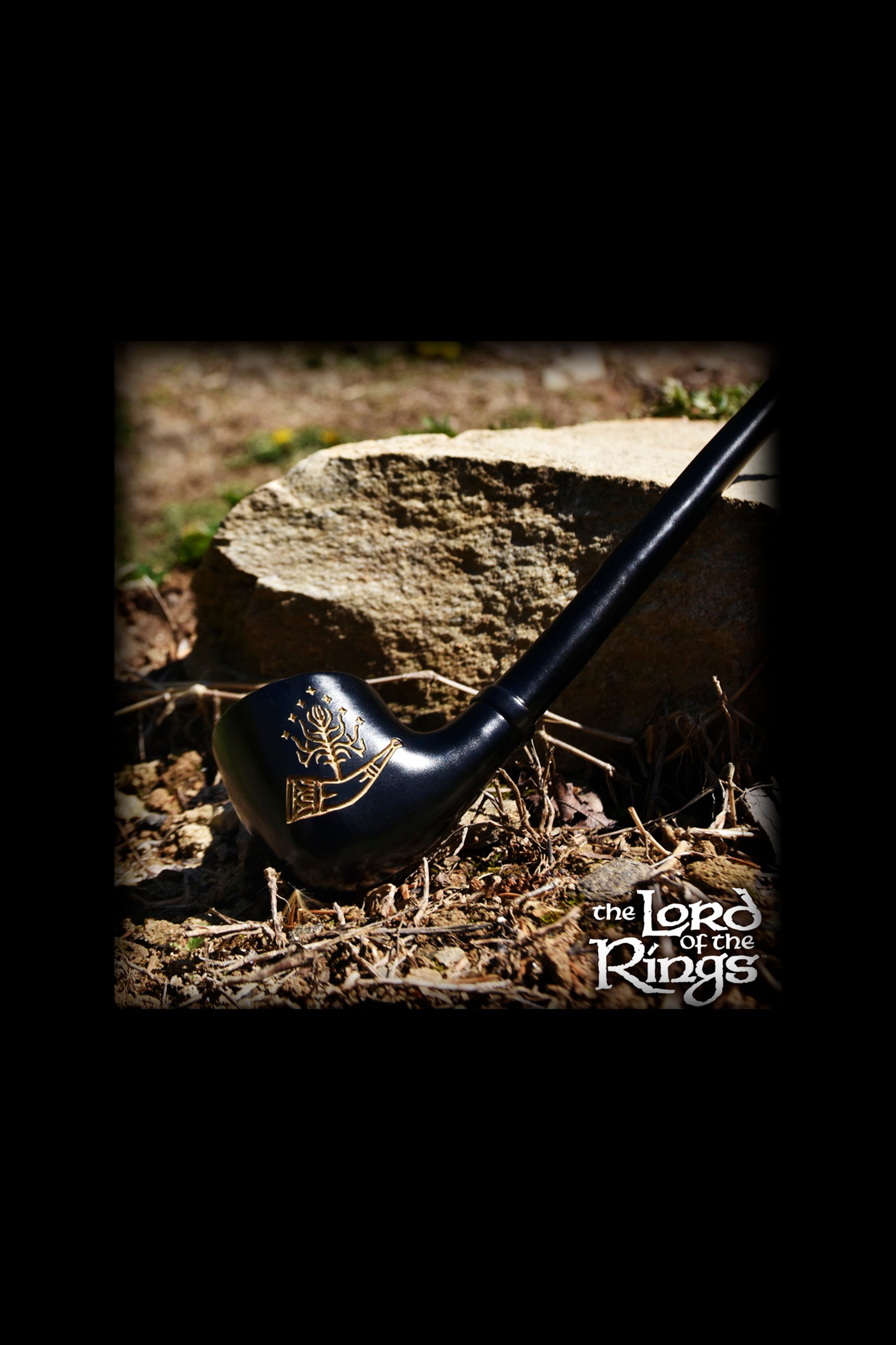 A black pipe with gold detailing rests on a tree stump surrounded by dry leaves and earth, evoking imagery from *The Lord of the Rings*.