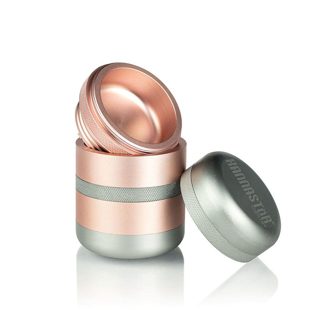 Angled close-up of the rose-gold and silver RYOT GR8TR Solid Body Grinder with its lid removed and resting beside the stacked halves on a white reflective surface