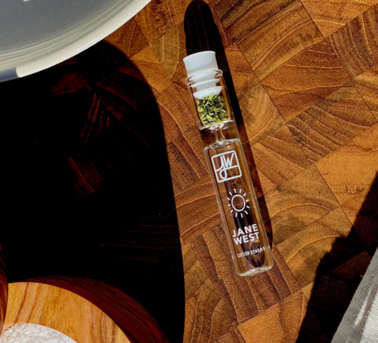 Glass vial filled with cannabis buds, labeled “Jane West” on a wooden surface.