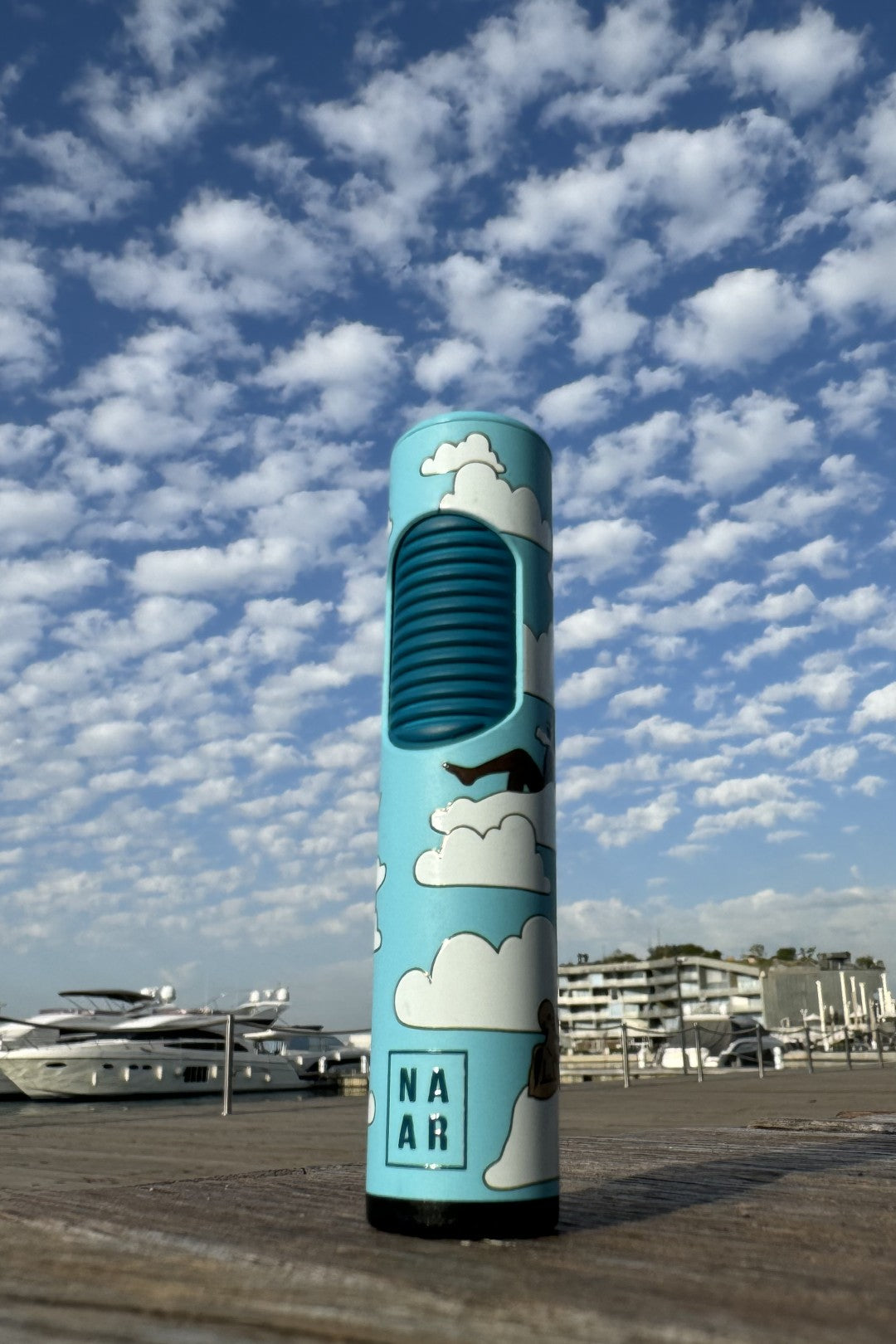 Blue, cylindrical cannabis vaporizing device branded "NAR" stands outdoors against a bright blue, cloudy sky, with yachts and buildings visible in the background.