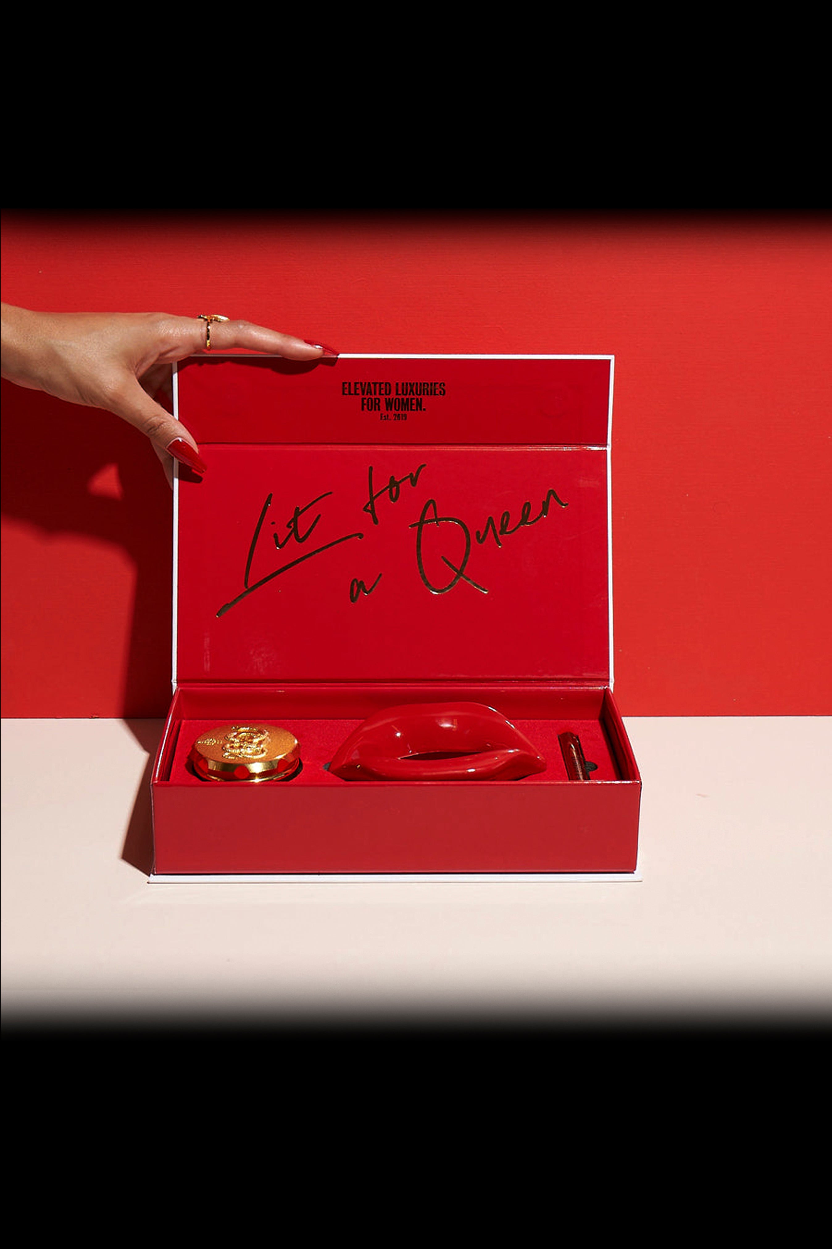 Red gift box with cannabis accessories and the handwritten message “Lit for a Queen” against a red background, with a hand reaching towards it.