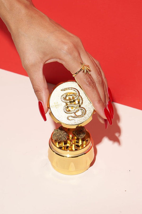 Gold grinder with cannabis visible, held by a hand with red nails, against a two-tone pink and red background.